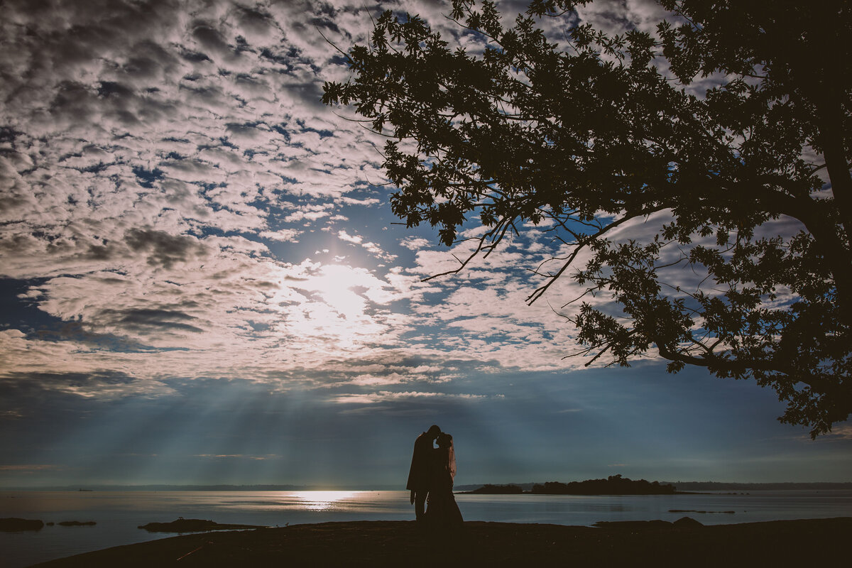 wide photo of the Sun breaking through the clouds with the bride and groom sharing a moment as a silhouette