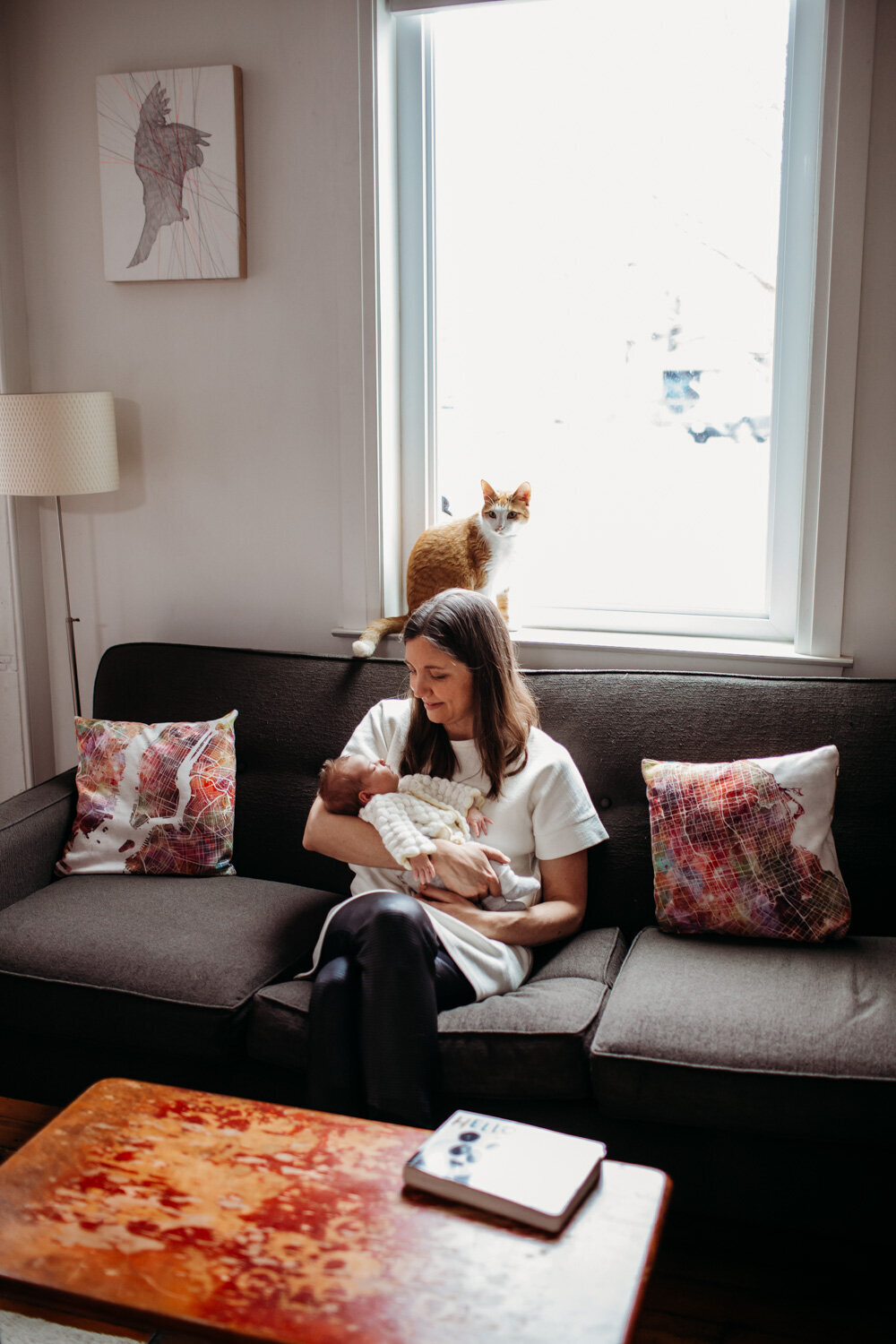 mom and newborn daughter sitting on couch with orange cat in window behind them