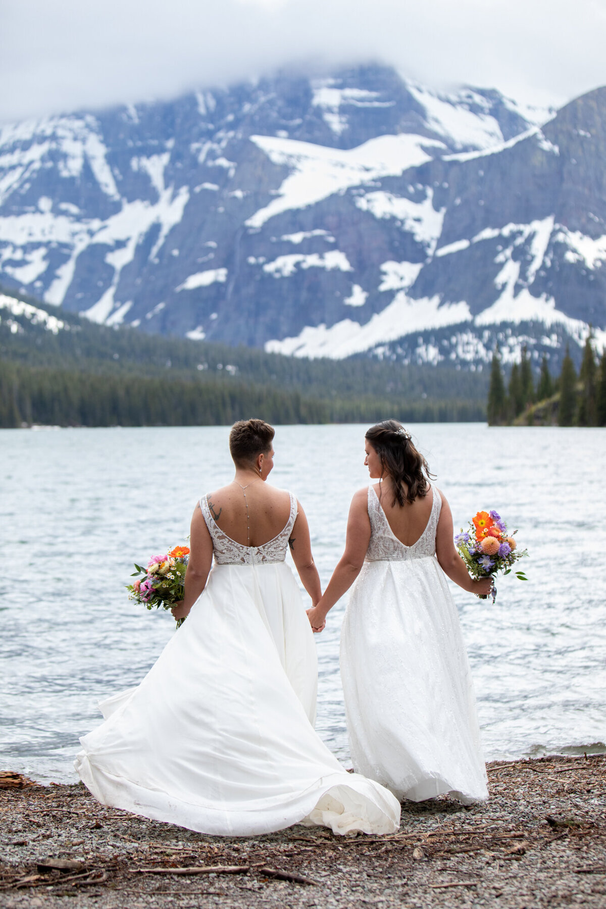 Two brides stand with colorful bouquets holding hands next to a lake in montana.
