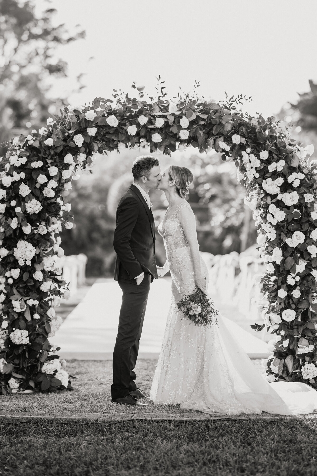 Bride and groom sharing a kiss under a floral arch, captured in a soft black and white image.