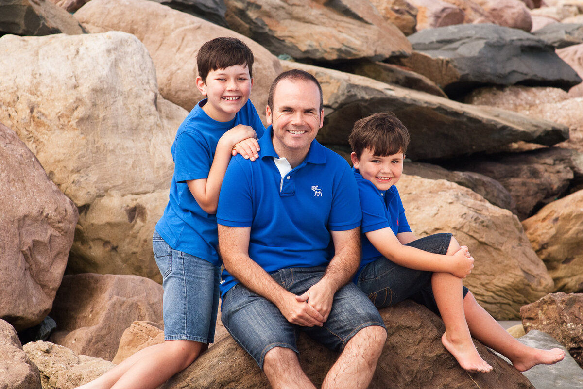 portrait of a father and two young sons sitting on beach rocks and smiling