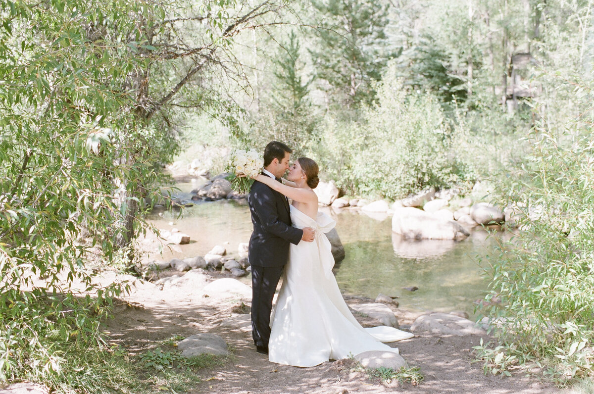 Bride and groom kissing under trees near a slow river