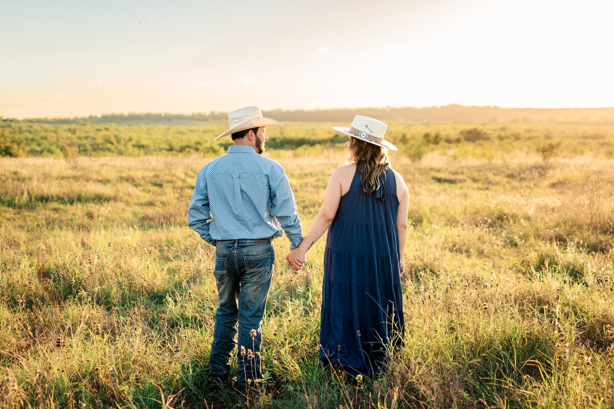 Engaged couple standing facing away from camera on  in open field during sunset engagement session at Harper Hills in New Braunfels, Texas.