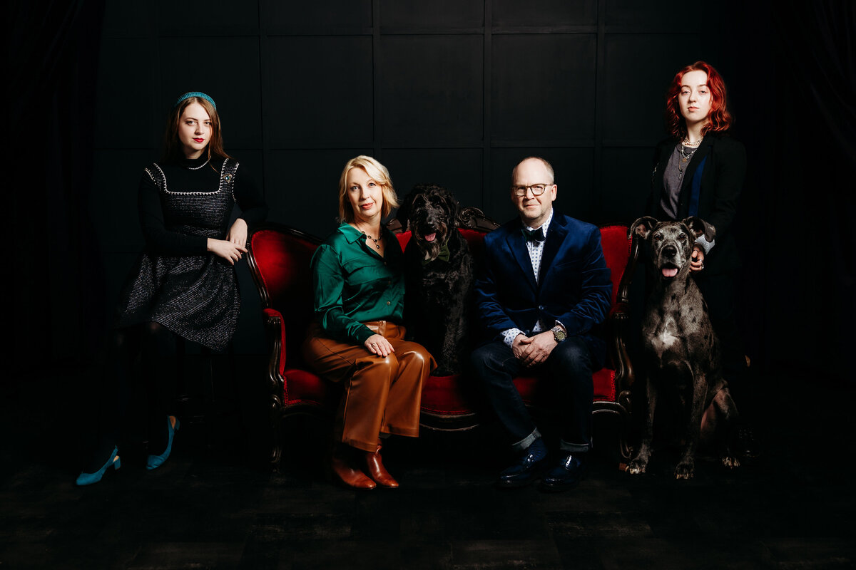 Serious studio family photo of 4 posing on red couch with two dogs. 