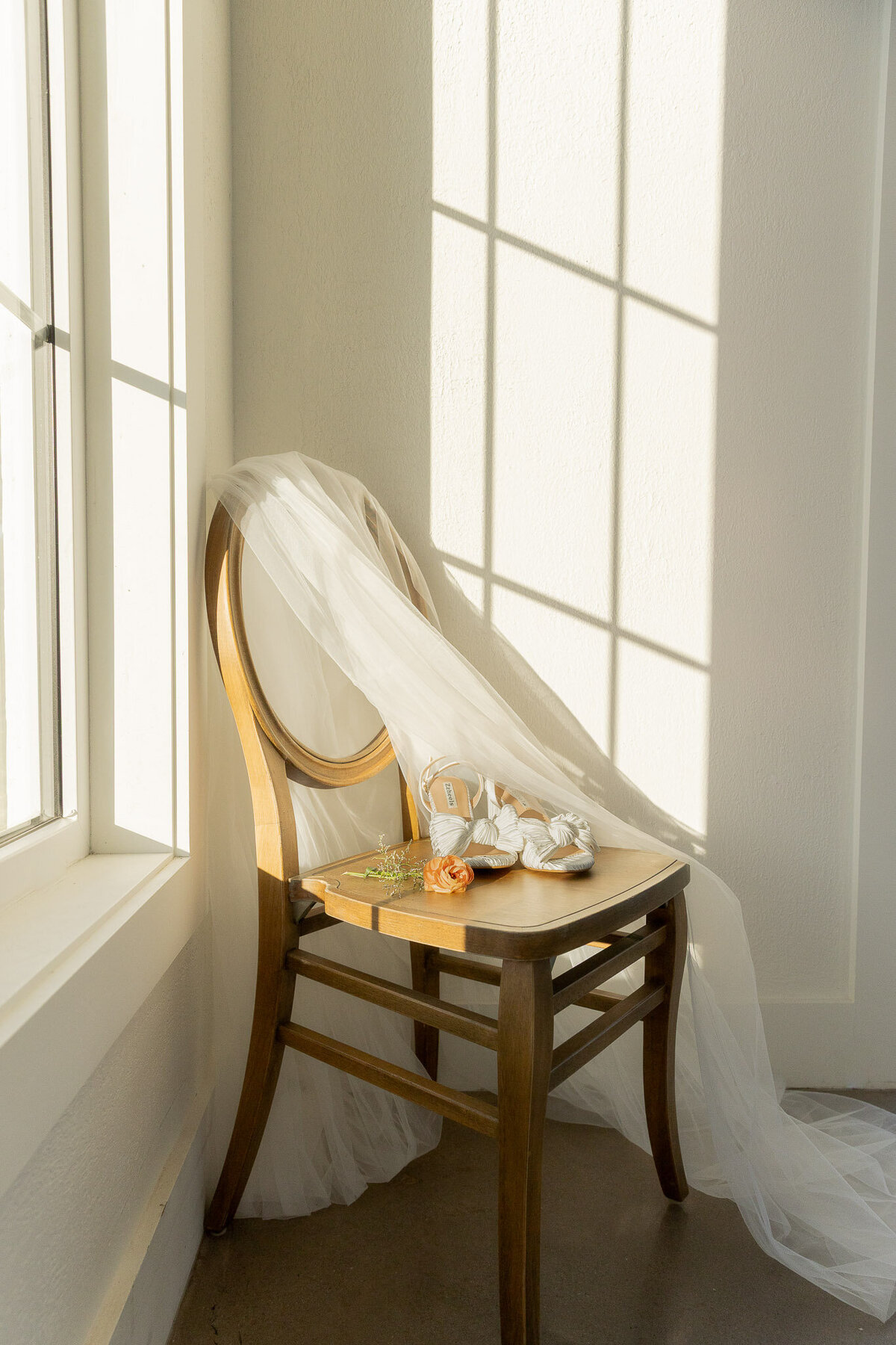 Detail shot of an orange flower and the bride's shoes and veil draped over a wooden chair inside of clean, white house.