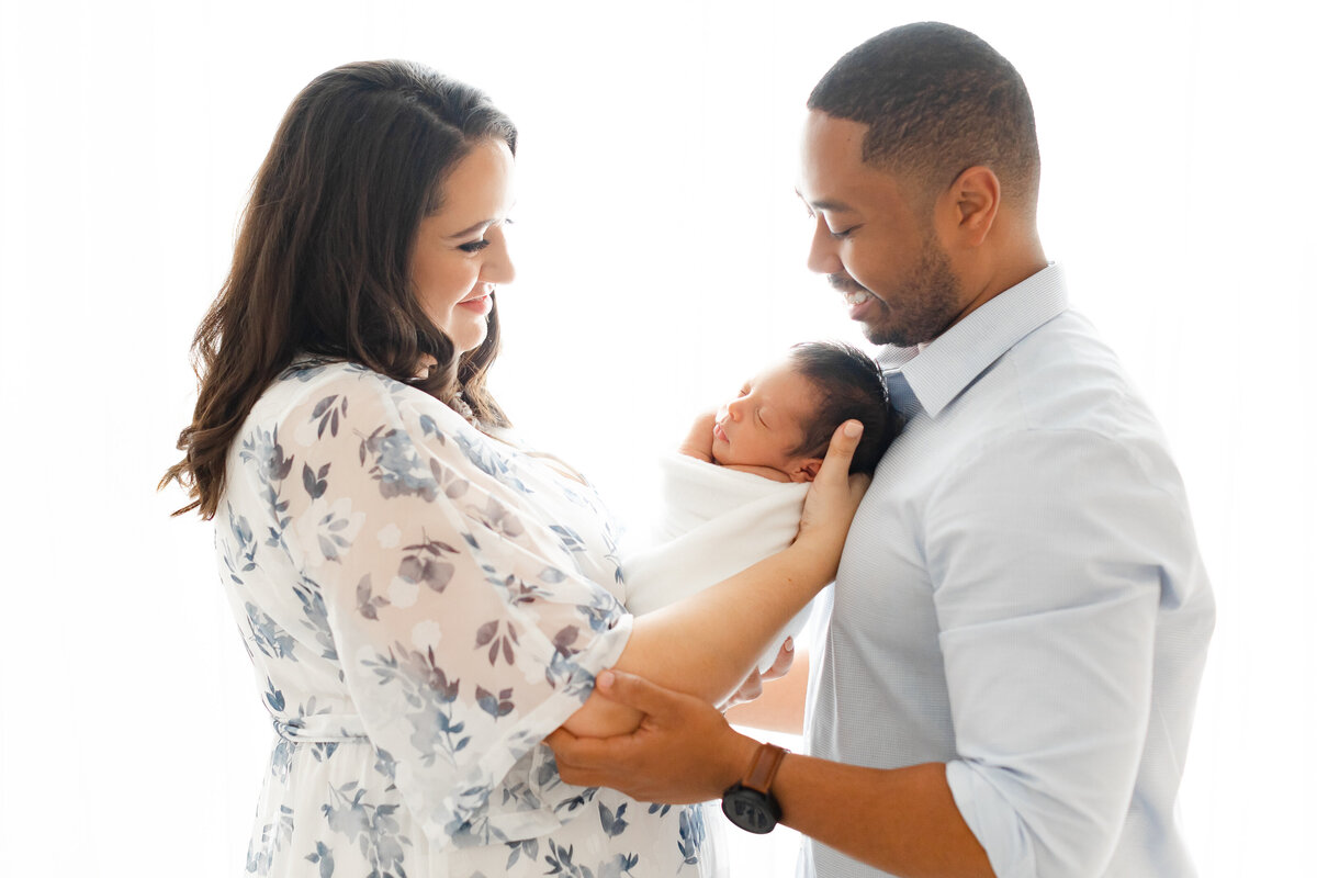 Two parents holding their newborn baby looking down at him smiling in front of a window at a Northern Virginia Family Photography session