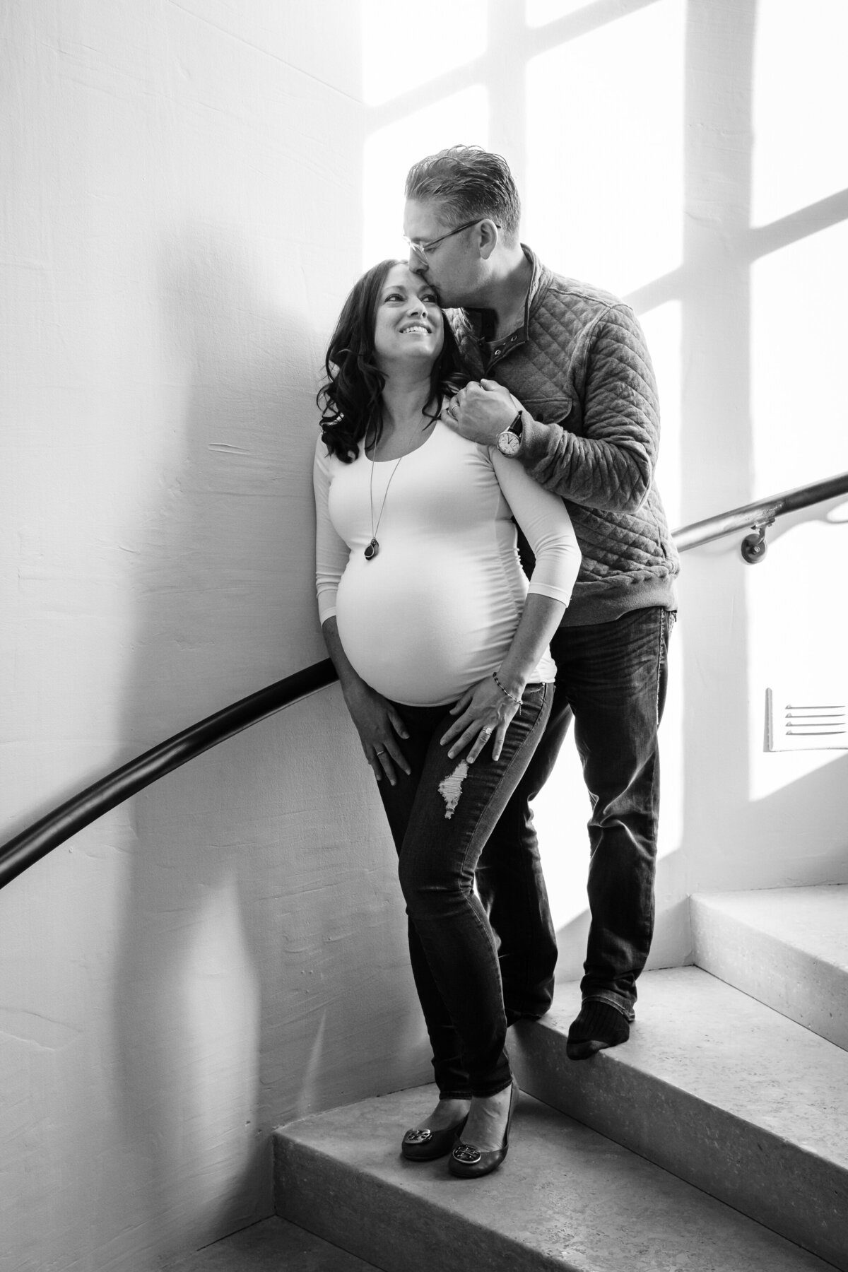 A couple stands on a staircase with soft light streaming through the windows. The man gently kisses the woman's head as he lovingly embraces her from behind, his hand on her pregnant belly.