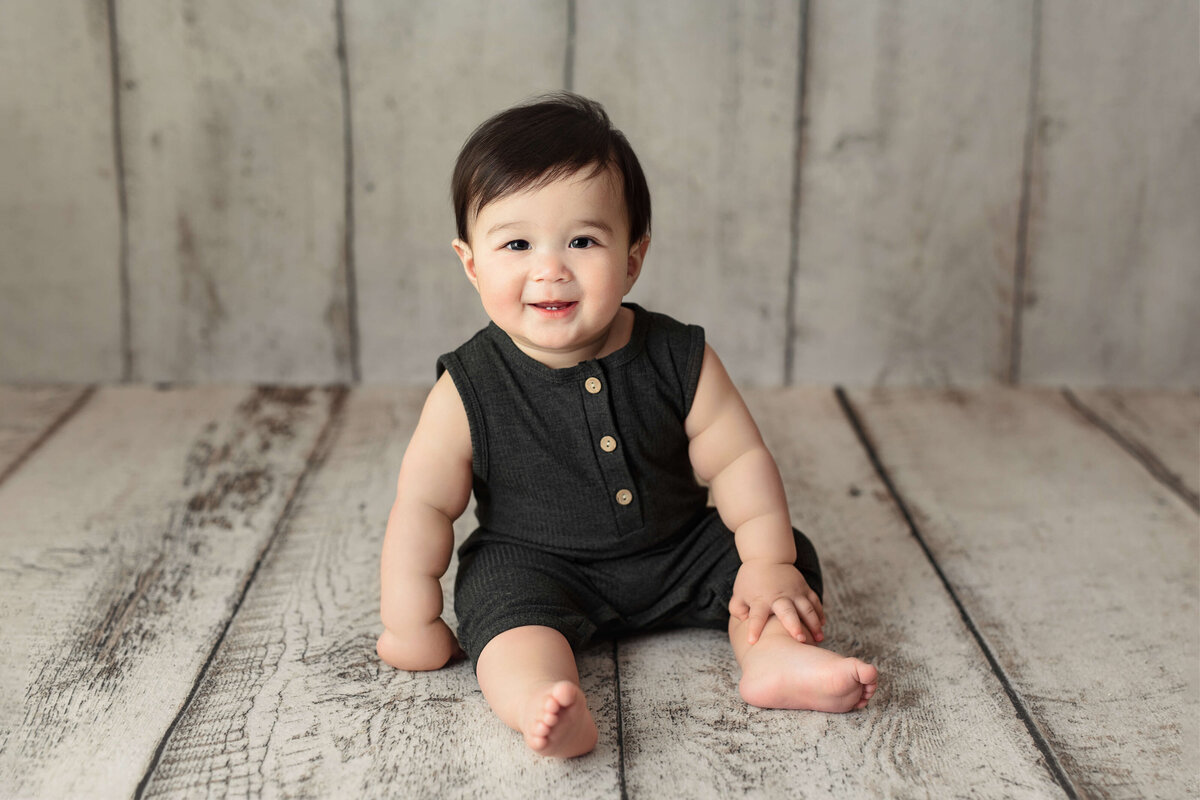 toddler in a dark grey onesie sitting on a white wood backgrpund at a baby photography session