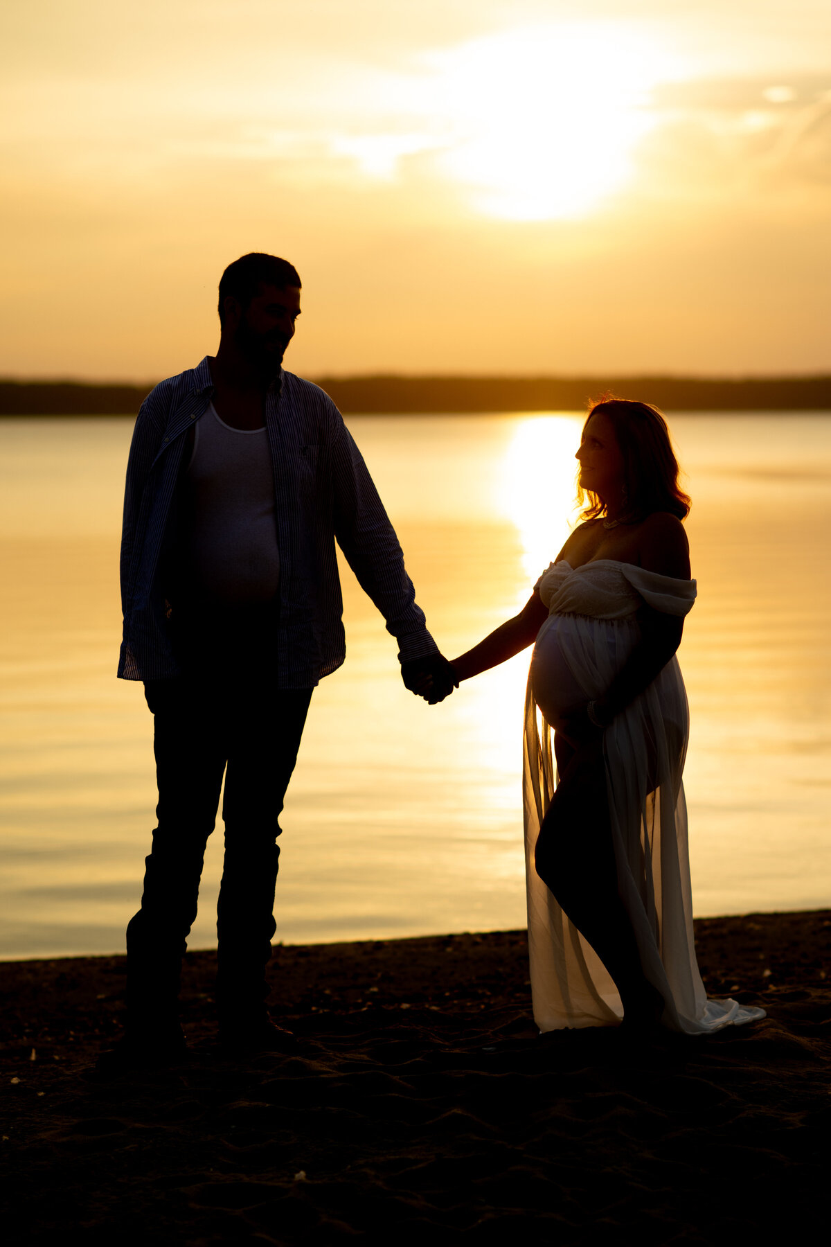 An expecting couple holds hands while standing in silhouette on a beach smiling at each other at sunset