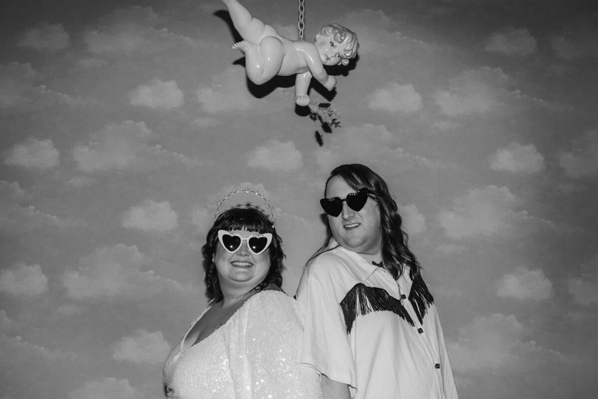 A black and white photo of two individuals wearing sunglasses, with a playful cupid decoration hanging above