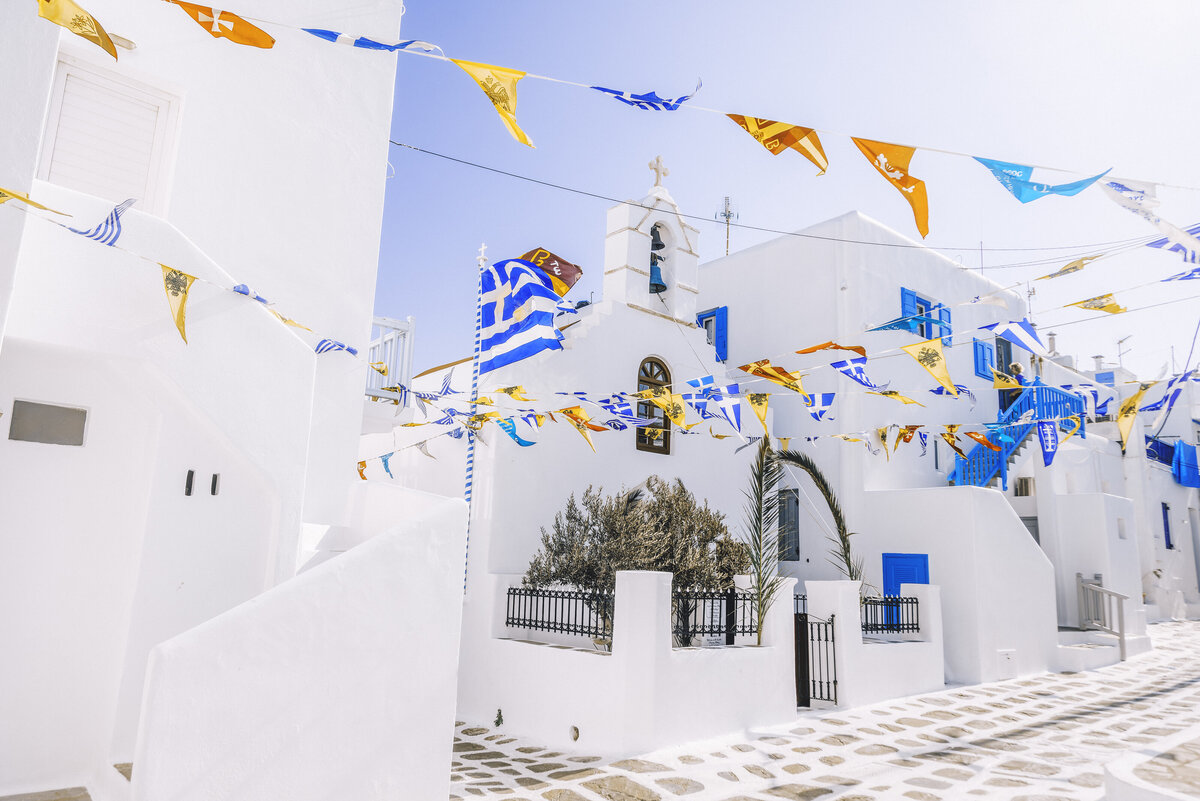 A village in Greece lined with white building and vibrant yellow and blue flags hanging overhead.