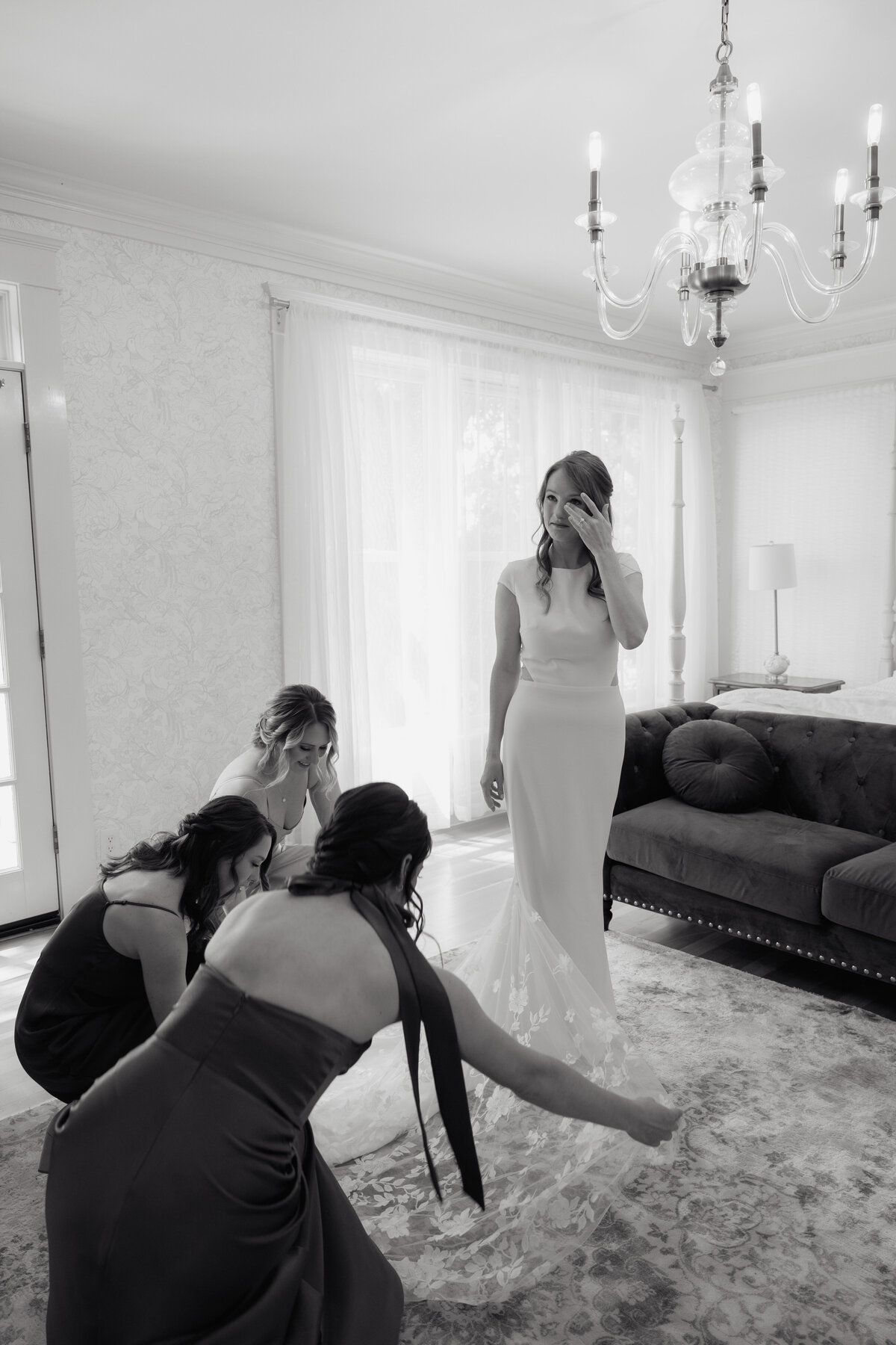 A bride wipes away tears as her bridesmaids adjust her dress, a heartfelt moment in a softly lit, elegant bridal suite.