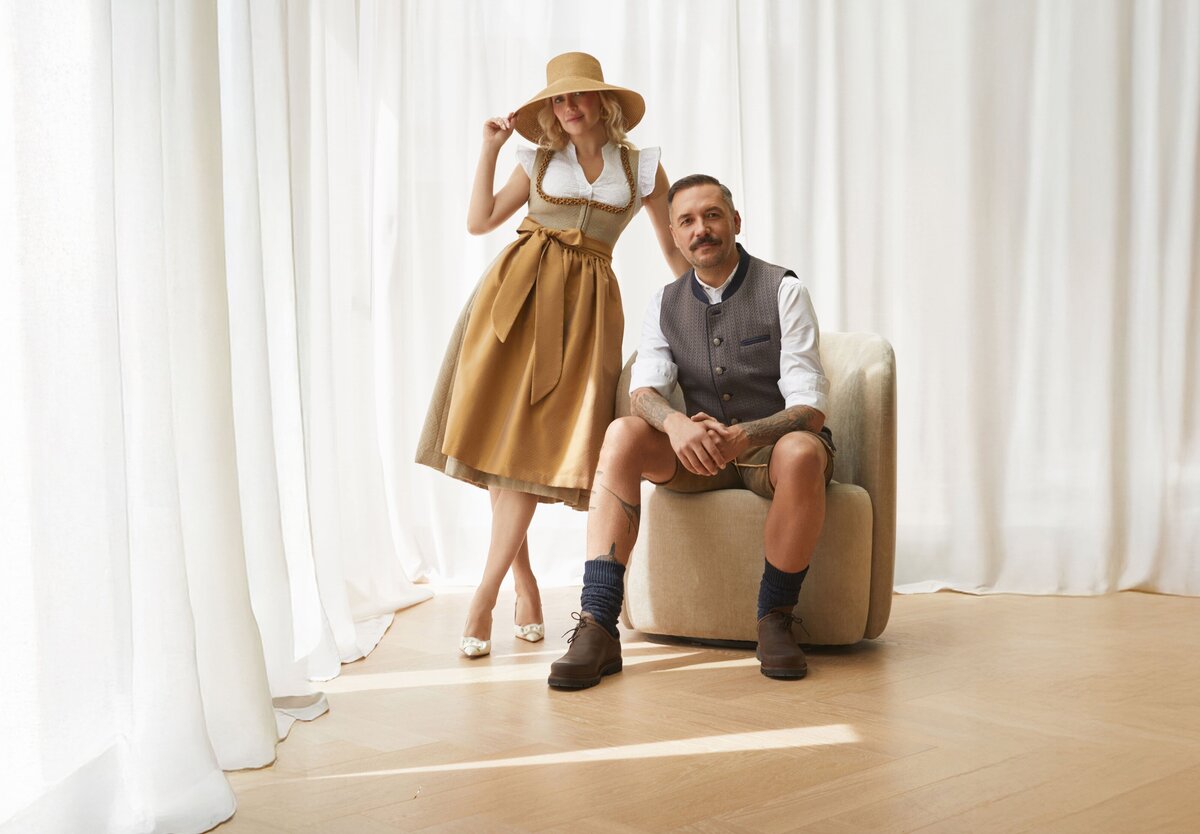 Stilvolles Trachtenpaar in moderner Designer-Tracht von Monaco Spatzl: Frau im gold-beigen Dirndl, Mann in Weste von Monaco Spatzl, Hirsch-Lederhose der Marke Schweigerts  – moderne bayerische Tracht aus München.