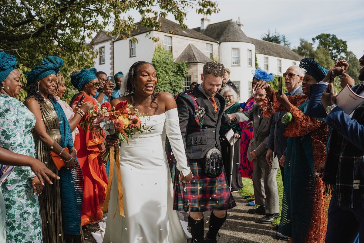 A bride and groom walk though two lines of wedding guests as they throw confetti above them. Everyone smiles and laughs as the bride and groom walk through them. Logie Country House is photographed in the background.