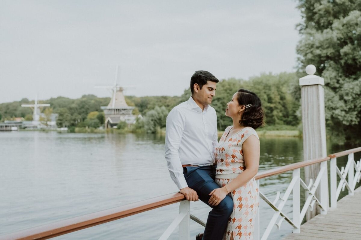 A posed photo of a couple on a wooden bridge, looking at each other with affection. The man, wearing a white button-down shirt and blue pants, sits on the railing. The woman, in a patterned, light-colored dress, stands next to him. In the background, there is a serene lake and two windmills.
