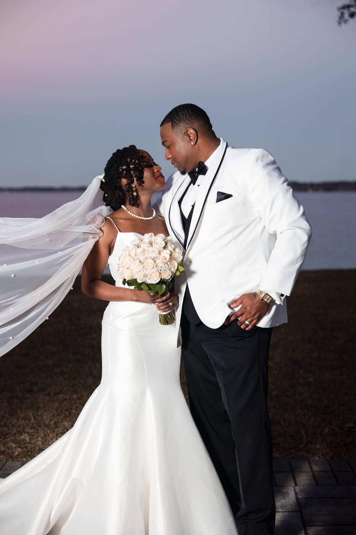 he bride and groom share a romantic gaze by the water at sunset during their wedding at Azaleana Manor in Orange Park, Florida. The bride’s veil flows in the breeze as she holds a bouquet of pale roses and stands beside her groom, who is dressed in a sharp white tuxedo jacket. The serene waterfront backdrop and soft evening light create a timeless and elegant moment. Captured by Heartful Films, a Jacksonville wedding photographer and videographer dedicated to documenting love with sophistication and heart.