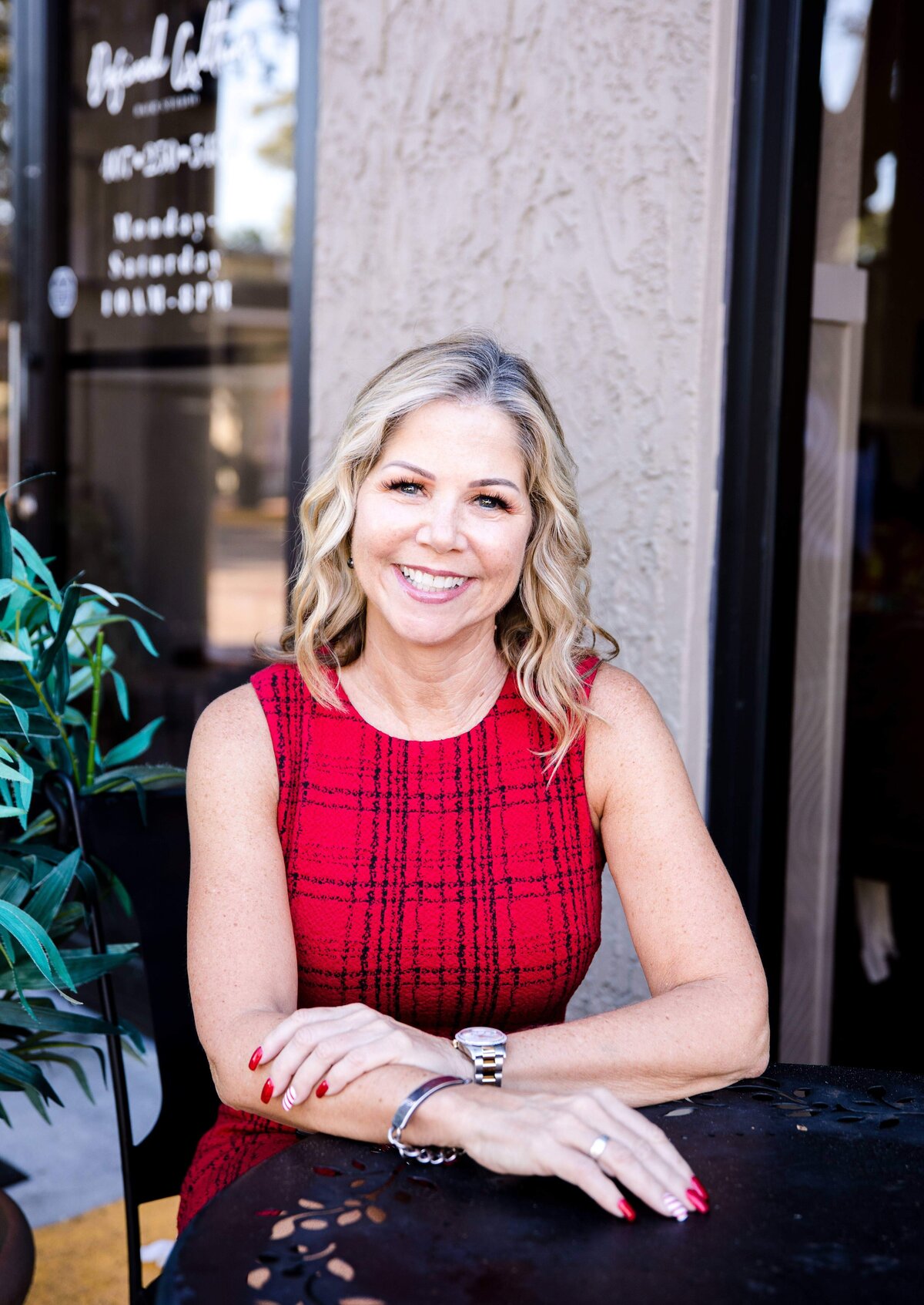 Orlando photographer photographs woman sitting at a cast-iron table outside of a local coffee shop with her hand on her forearm resting her elbows on the table smiling while in a red dress for brand photography in Orlando