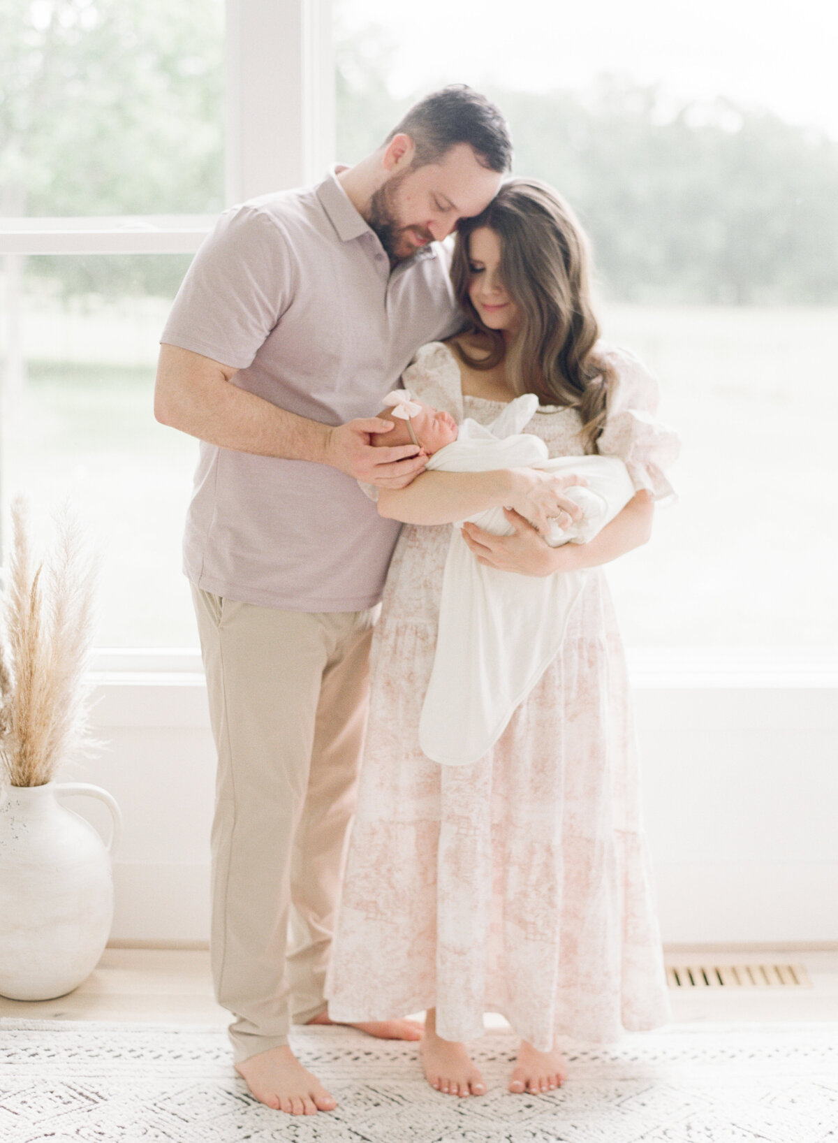 Mom and dad snuggle newborn daughter in front of giant window during the Raleigh newborn photography session. Photographed by Raleigh newborn photographer A.J. Dunlap Photography.