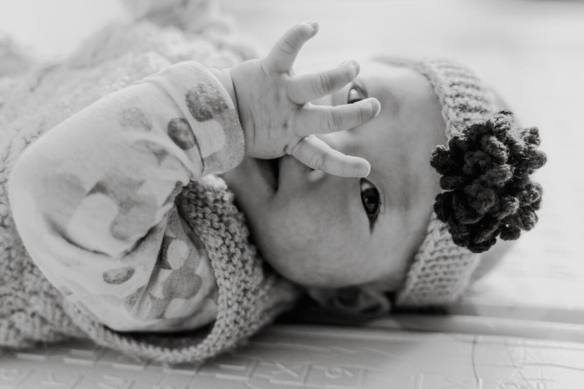 Black-and-white close-up of a baby wearing a knitted hat with a flower detail, sucking on their hand while lying down.