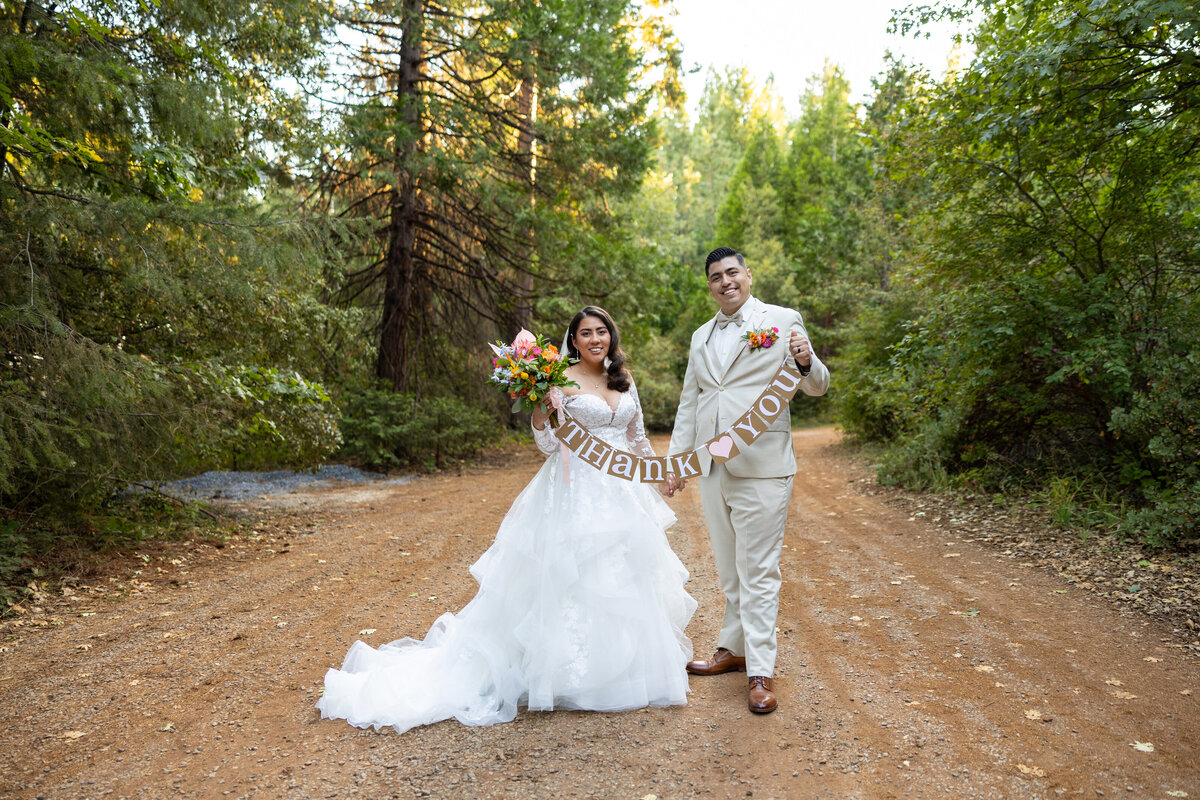 Bride and groom stand on a path lined by trees at their wedding and hold out a thank you sign, for a photo that will be used in a thank you card later for their guests. Photo captured by Philippe studio pro, Sacramento wedding photographer.