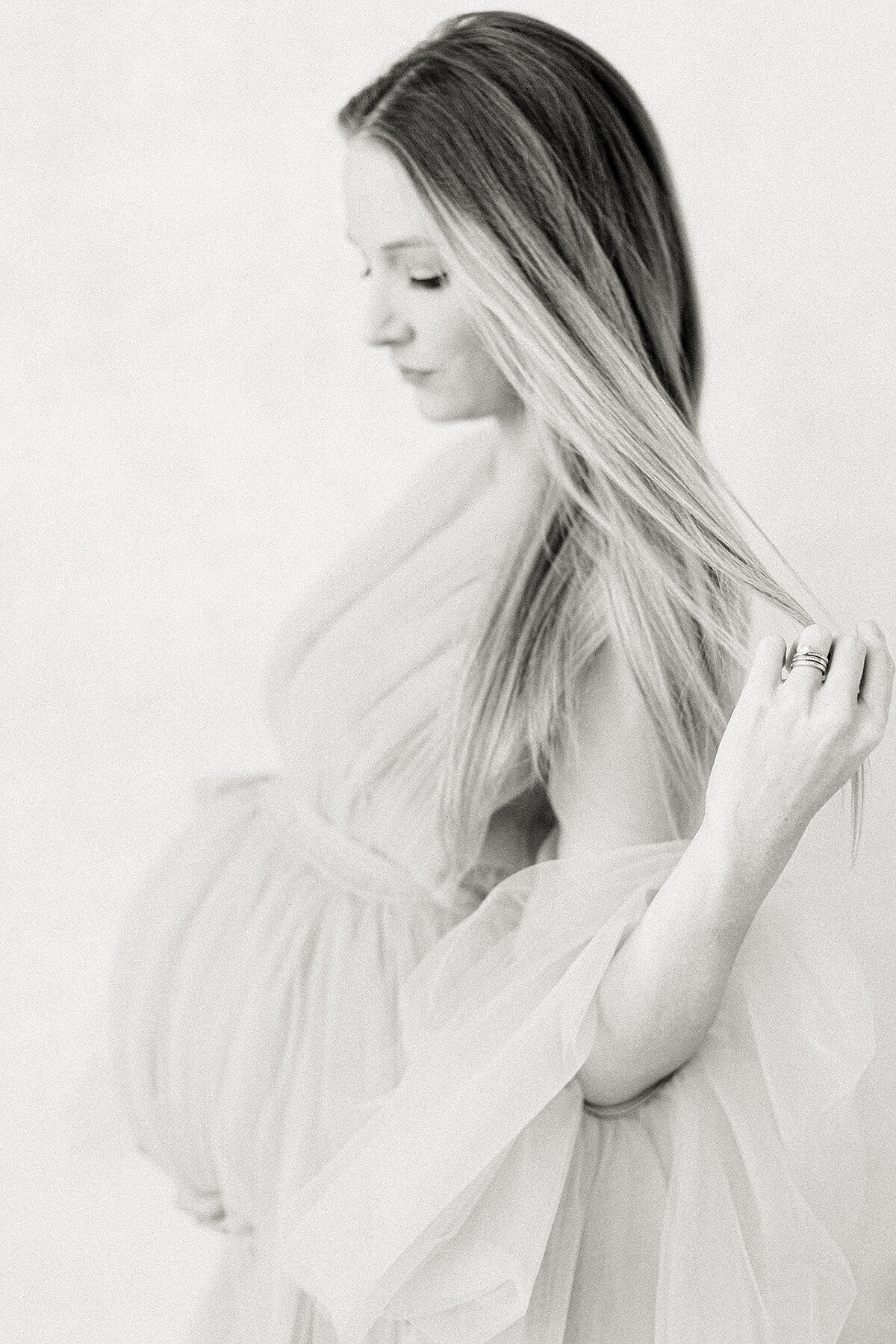 Black and white in studio maternity portrait of a mother wearing a tulle maternity gown while she is holding her hair back away from her face.