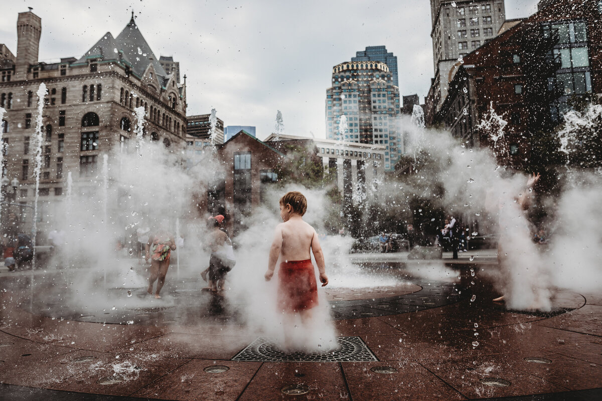 a boy in red shorts stands over a grate at a splash park in Boston Seaport with steam and water coming out. 