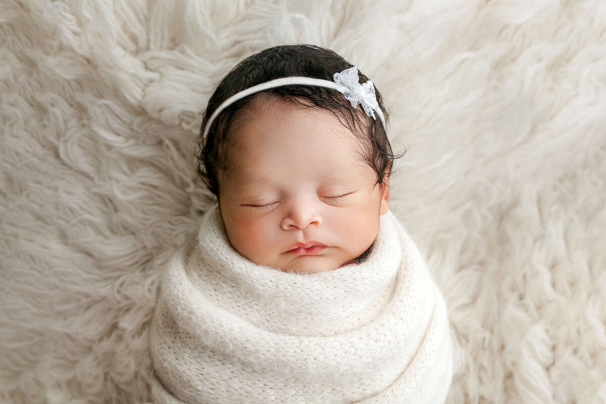 newborn baby girl in a white knot swaddle f on a white fur rug with matching headband at a newborn photo shoot in Northern VA