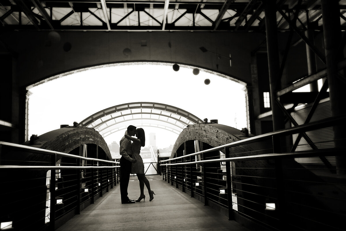 A couple kissing while standing on a walkway under a bridge 