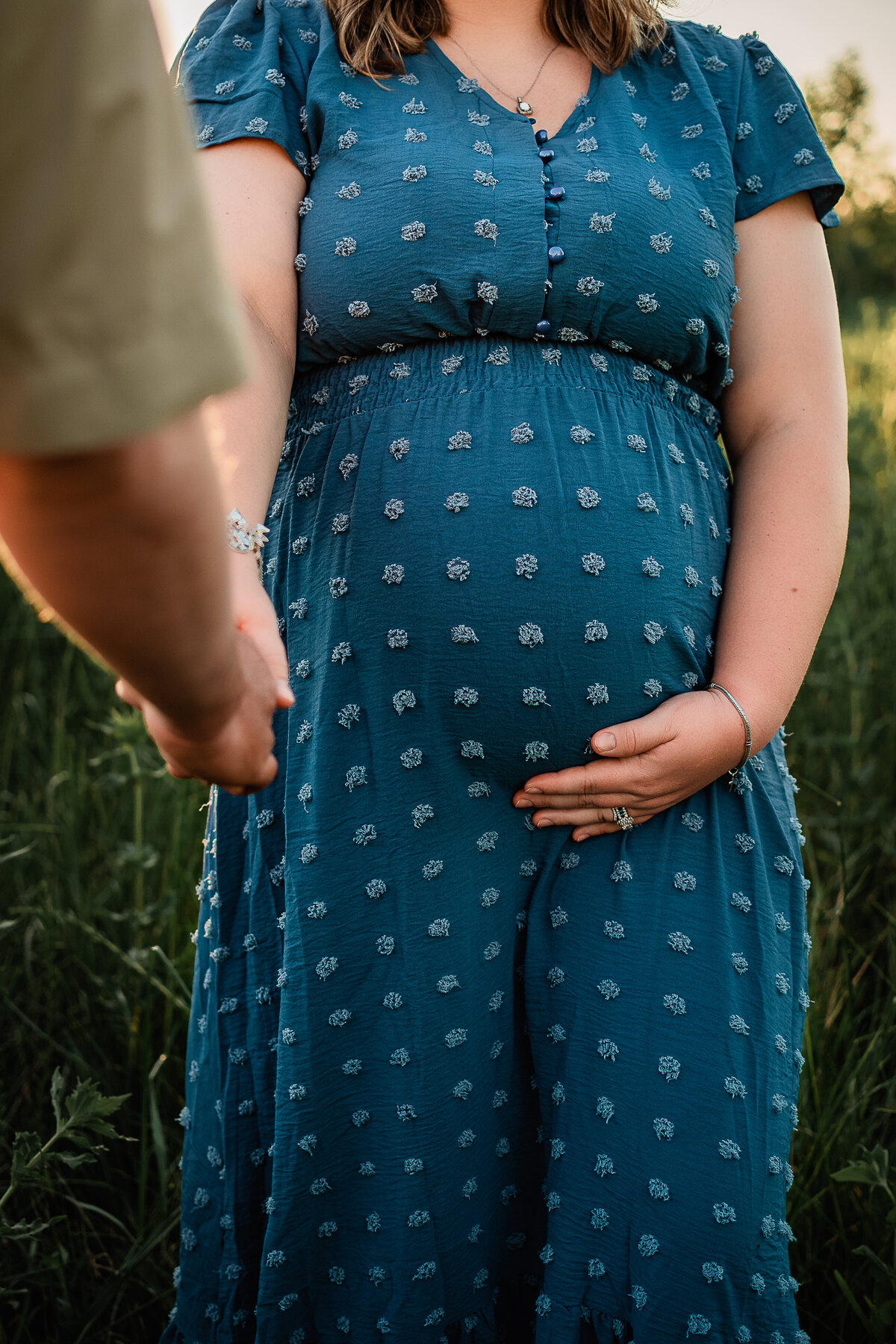 A woman in a blue dress holds her husband's hand and her belly for a close up detail shot.
