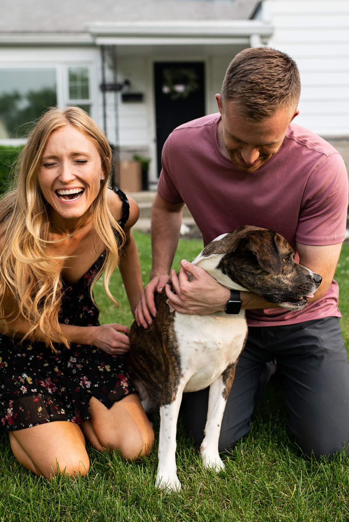 Couple sits with dog and laugh during engagement session at their home