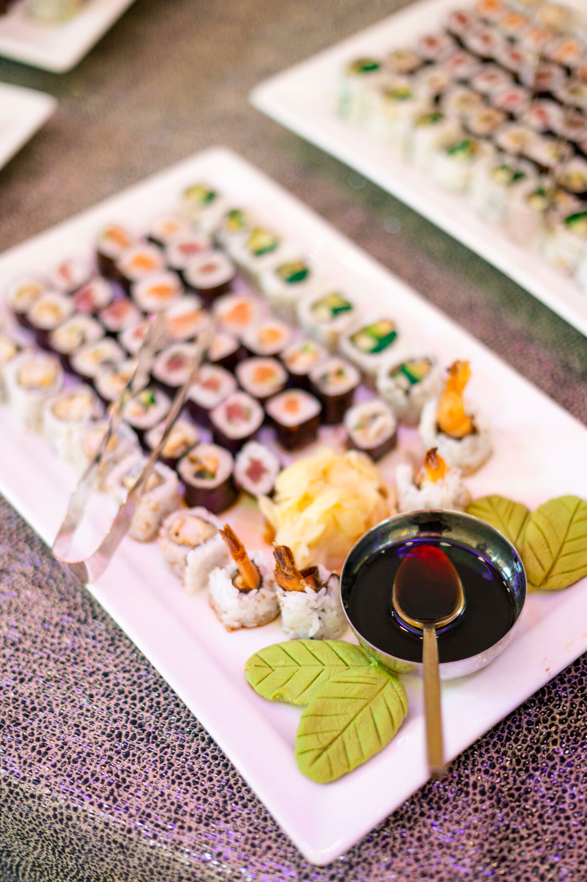 A rectangular platter of assorted sushi rolls, including shrimp tempura, alongside a small dish of soy sauce with a spoon. Ginger is piled in the center, and two green leaf decorations adorn the plate. Sushi tongs are placed nearby.