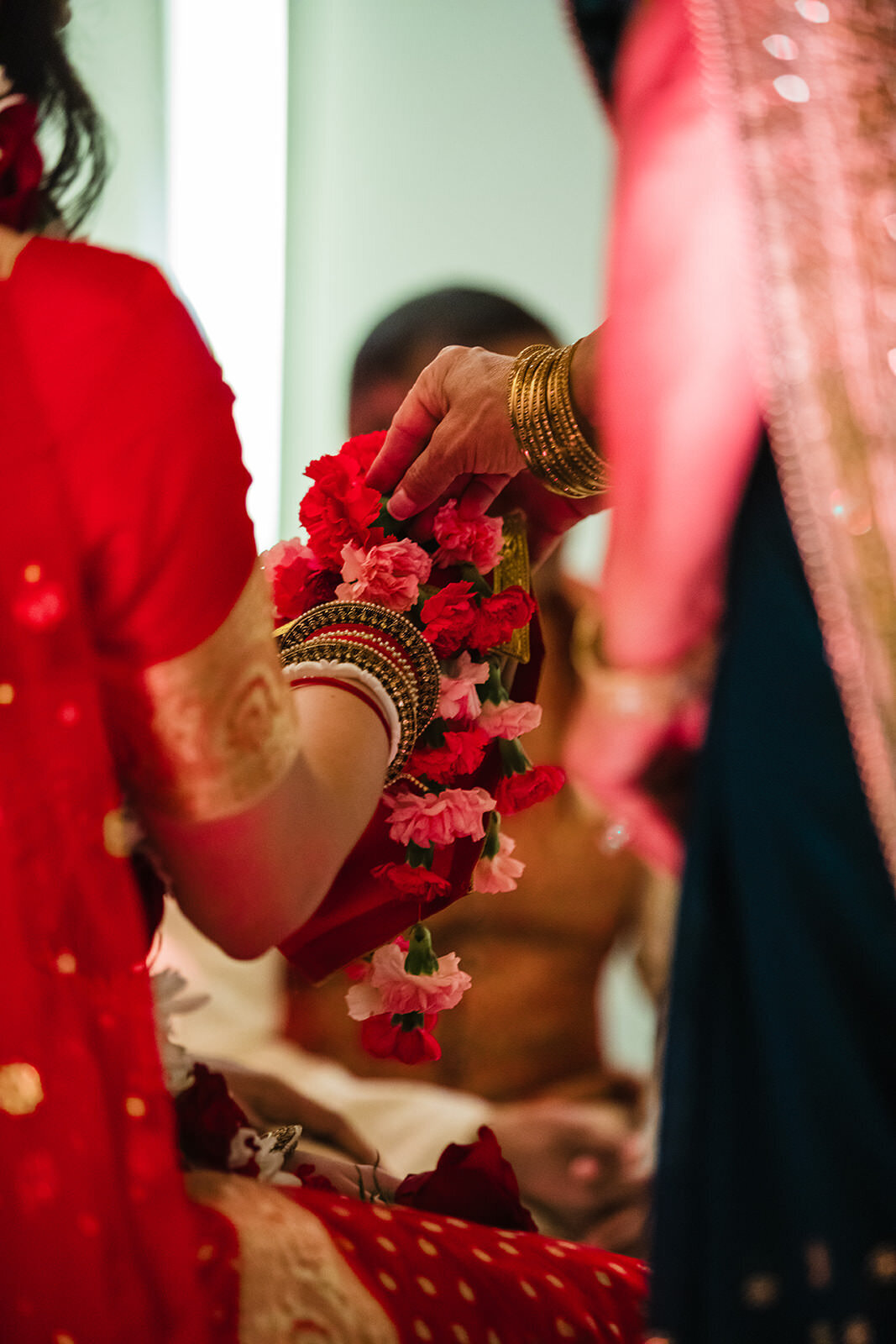 A close-up of a hand placing floral garlands on the wrist of a bride in red attire, with focus on the traditional Indian wedding bangles.