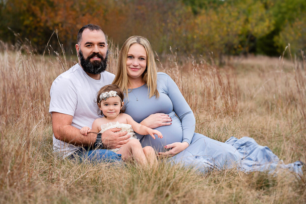 Expectant mom, dad, and toddler are sitting in a field for maternity photographs. Mom is leading against dad and the little girl is sitting on dad's lap. All are looking at the camera. Captured by Main Line photographer Liza Nicole.