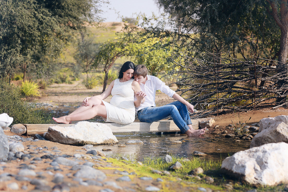 Couple cuddling while sitting by the water in nature during the pregnancy photo session by Calgary Family Photographer.