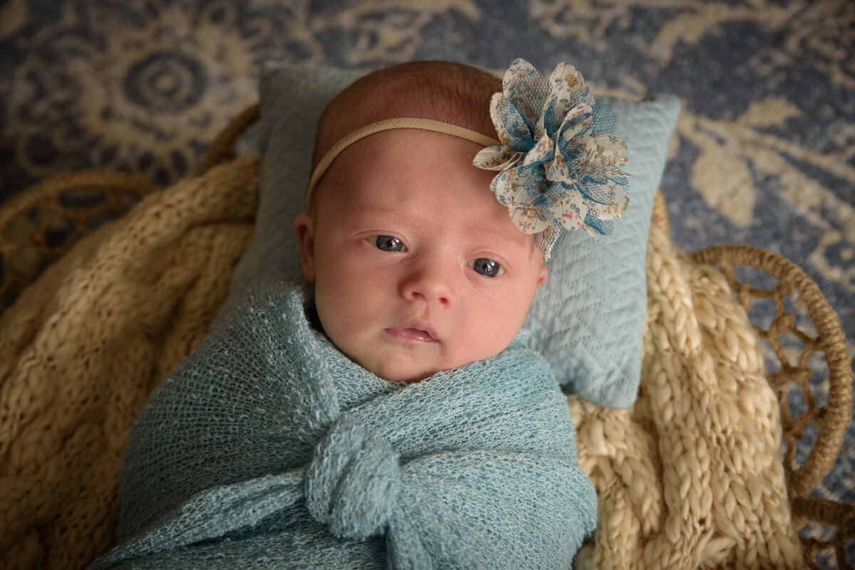 Newborn baby girl in light blue wrap with a cream and blue bow posed in a basket in her home in Green Bay, Wisconsin