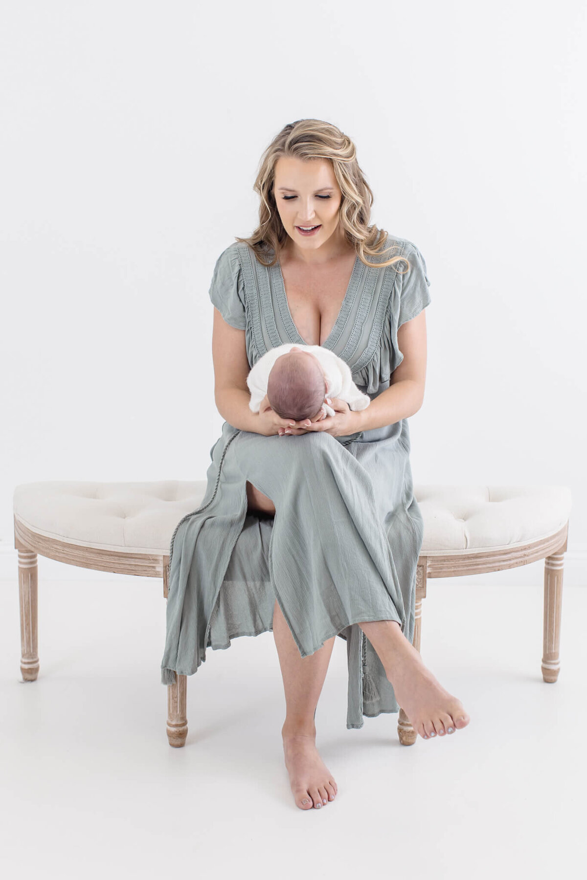 A woman in a light blue dress sitting on a cream-colored bench holds a newborn baby wrapped in white. She smiles down at the baby in a serene, well-lit setting.