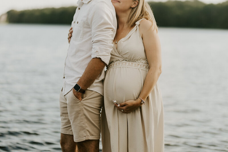 Couple collé l'un à l'autre devant un lac entouré d'une forêt. La femme enceinte tient son ventre avec sa main. Shooting photo grossesse en Vendée.