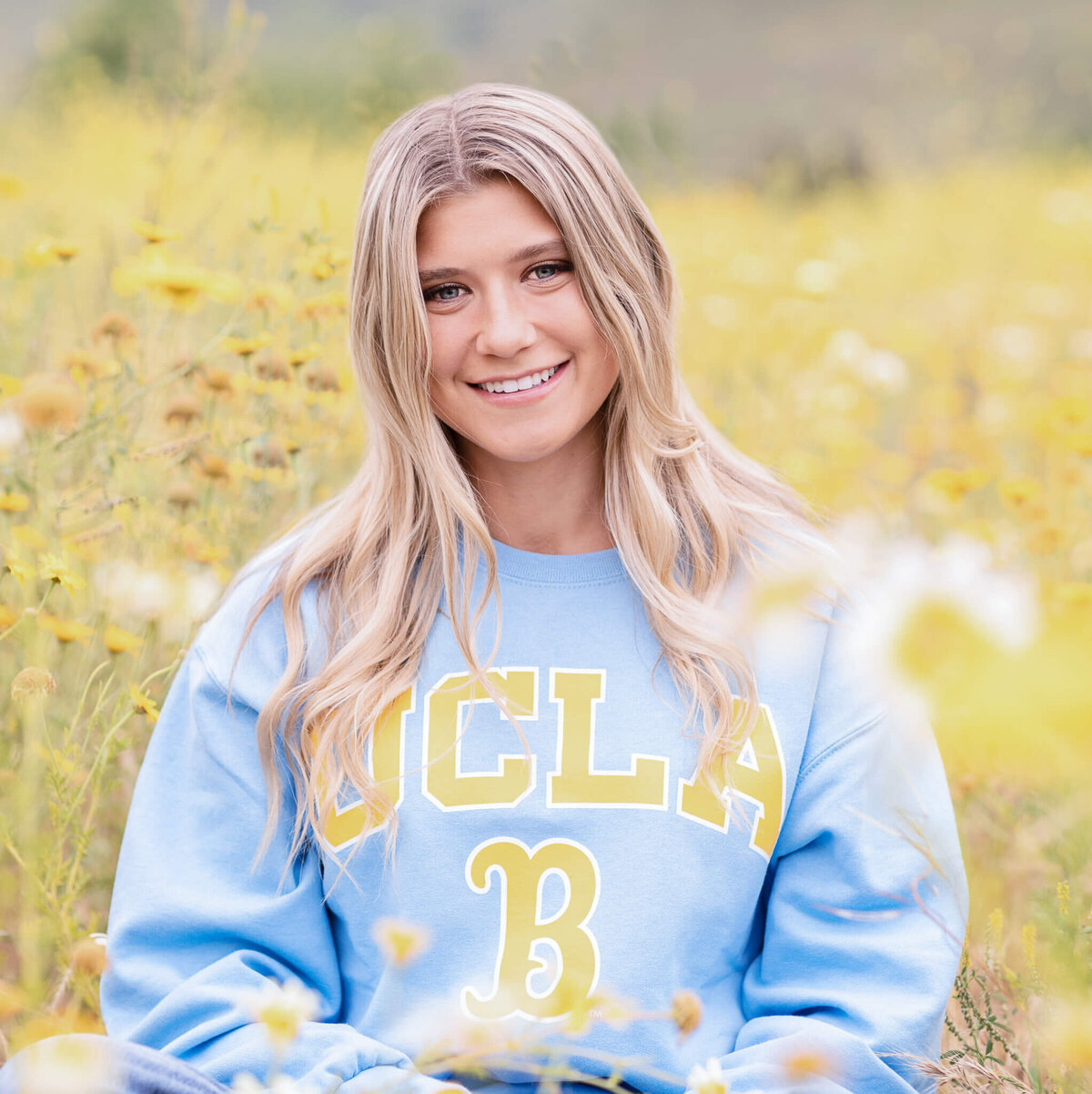 Smiling person with long blonde hair wearing a blue UCLA sweatshirt sits in a field of yellow flowers.