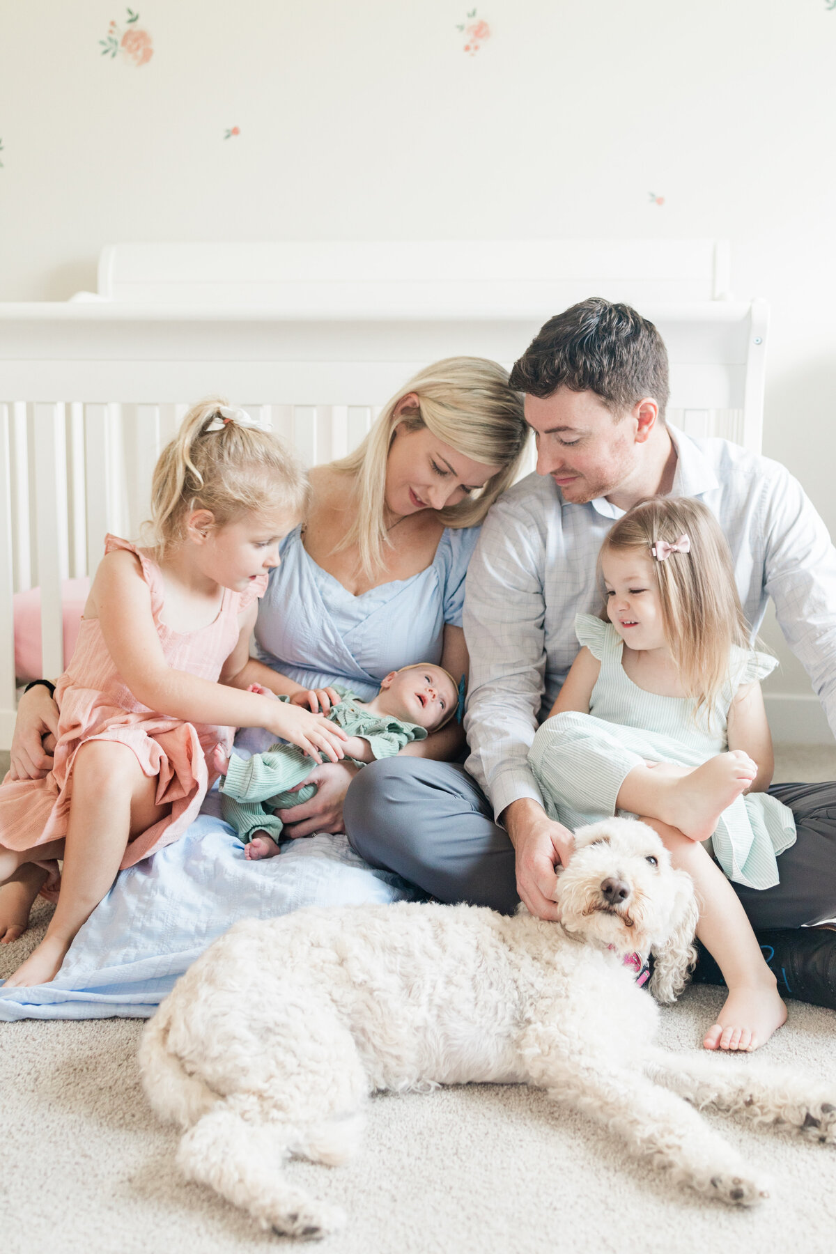 Family cuddled in front of a crib with a newborn and puppy