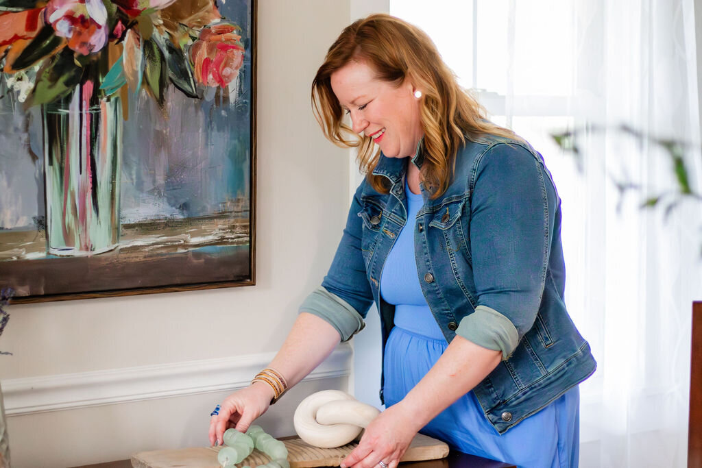 A smiling woman with shoulder-length reddish-brown hair, wearing a light blue dress and a denim jacket, is arranging small, light green decorative objects on a wooden surface. A larger, white decorative knot sits beside them. Behind her is a white wall with a framed painting of a vibrant floral arrangement in a tall vase. A window with white curtains is visible to her right, providing soft natural light.