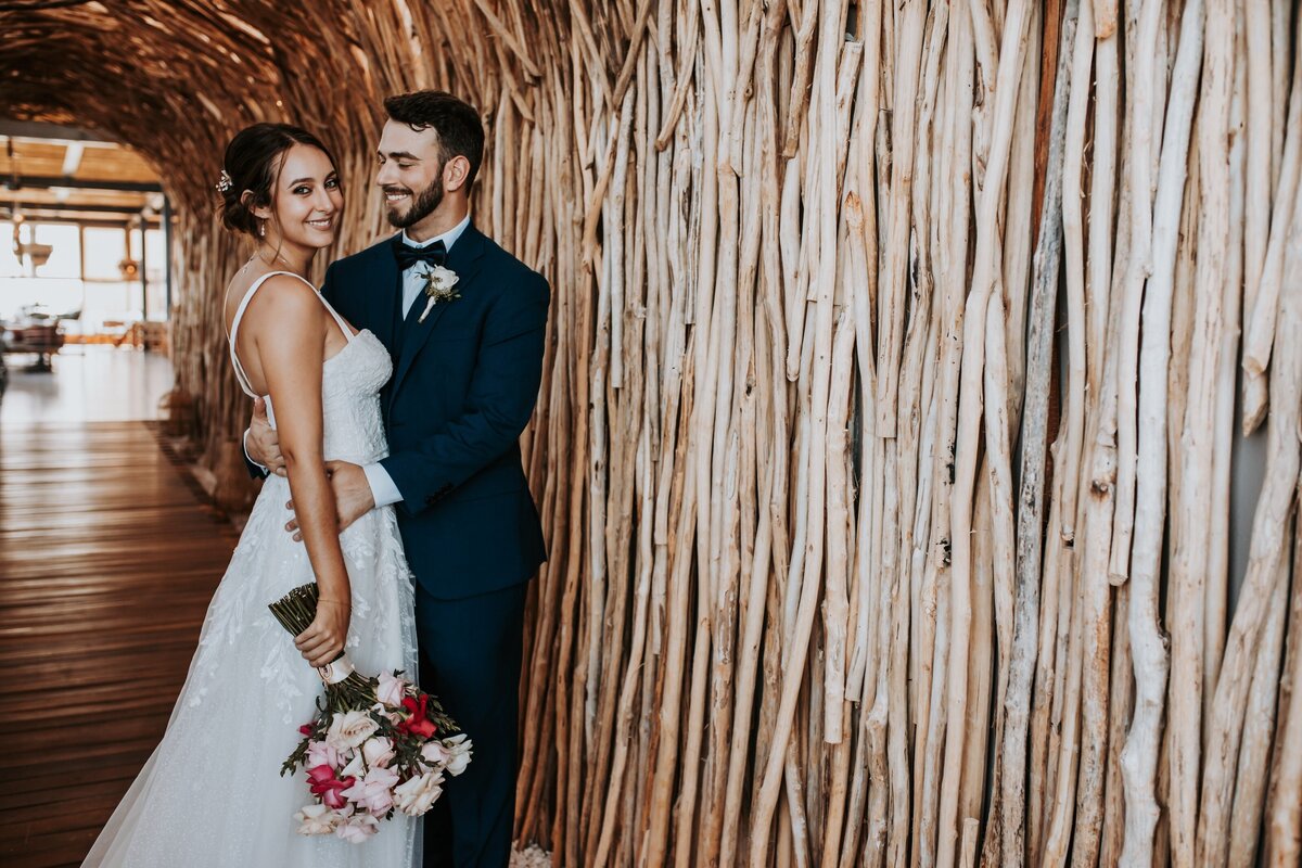 A bride and groom pose together inside a venue with a textured wooden wall. The bride wears a white gown and holds a bouquet of pink and white flowers, while the groom is dressed in a navy blue suit and looks at her with a smile.