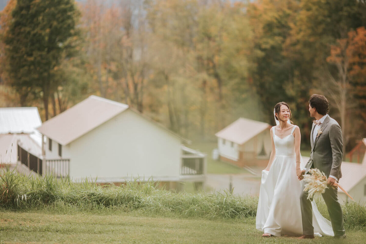 A couple walks hand in hand outdoors, surrounded by autumn foliage and rustic buildings