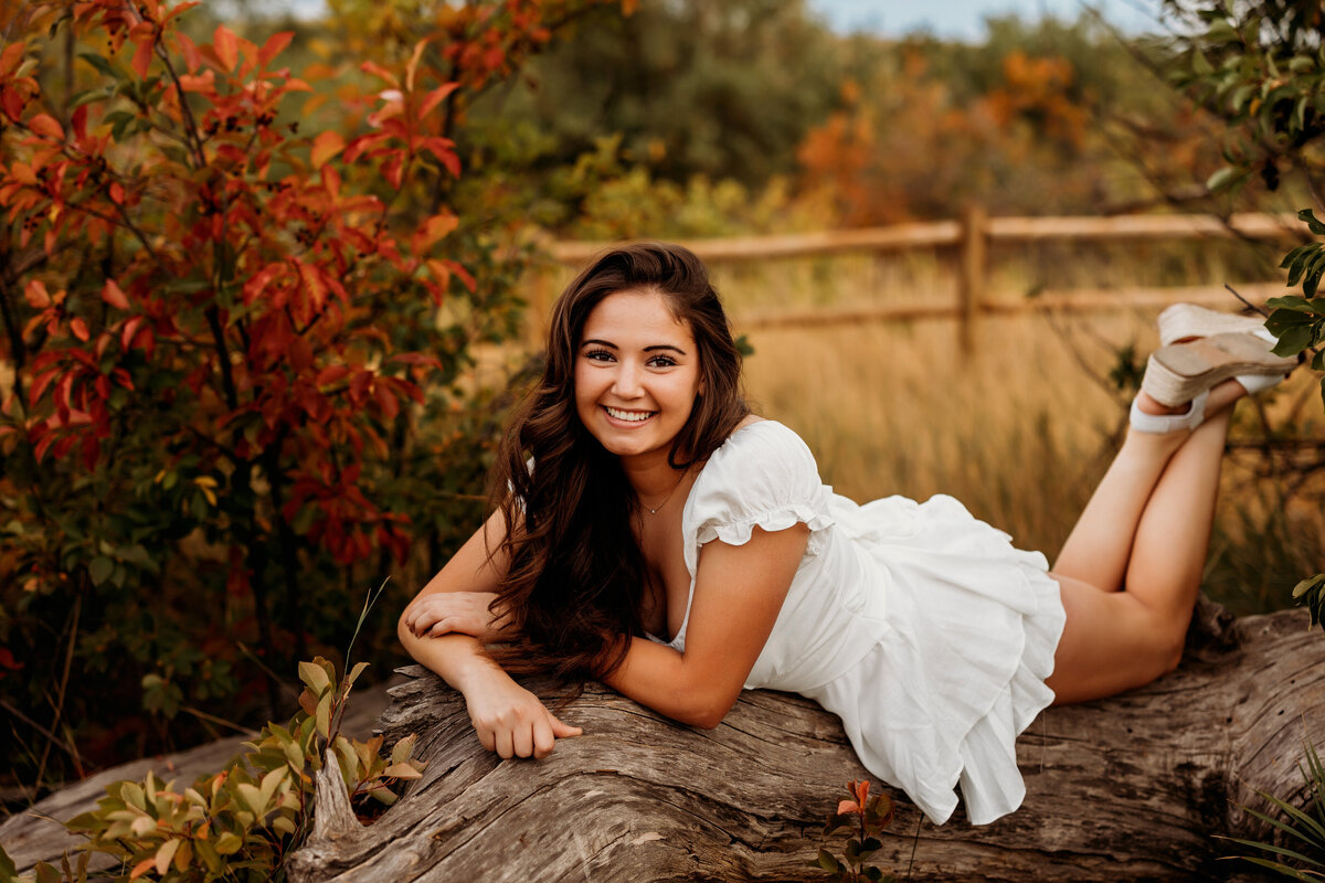 High school senior girl wearing a white dress lays on her stomach on top of a tree trunk in the middle of fall