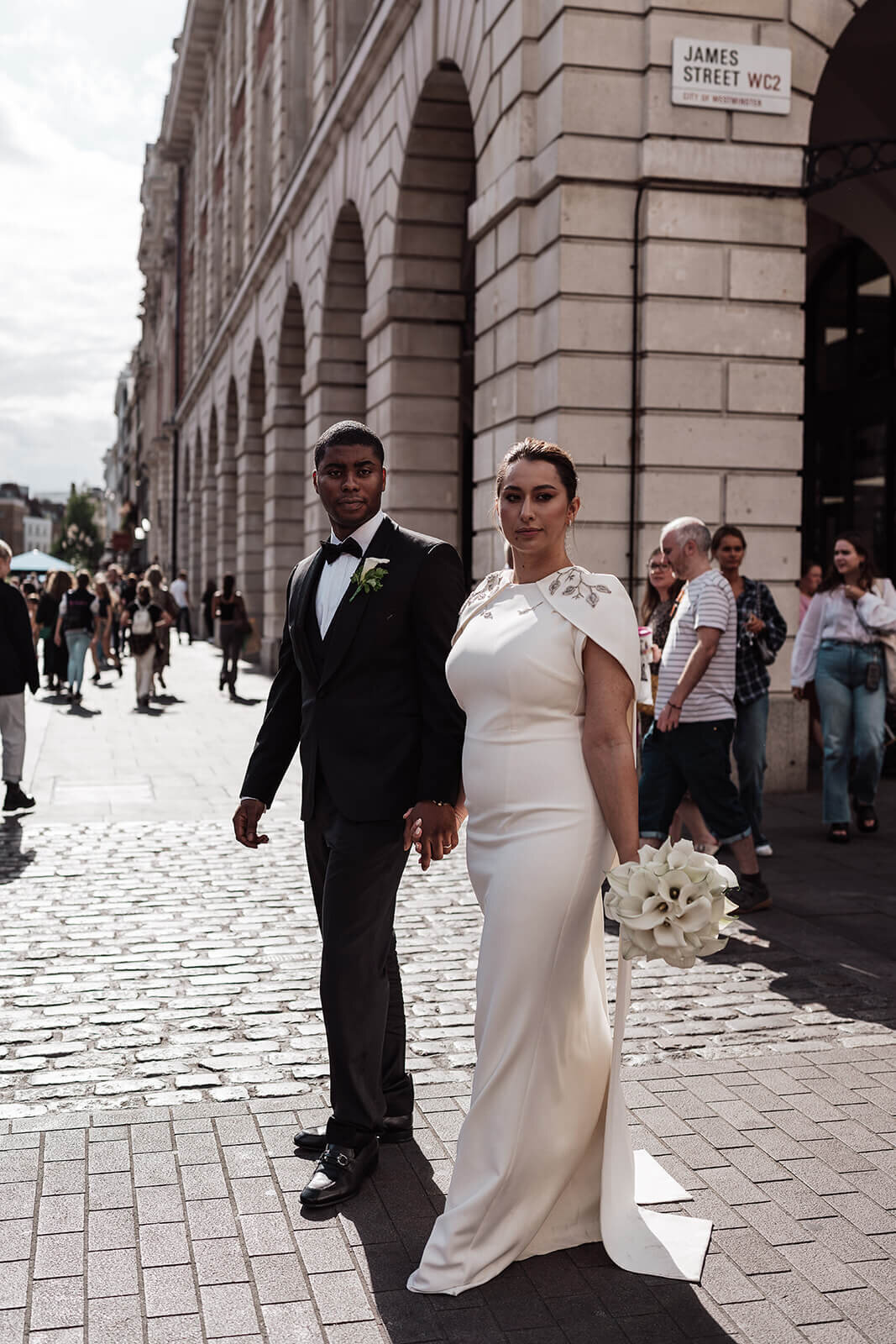 bride and groom hold hands and walk through the streets of covent garden london on their wedding day as people mill around them