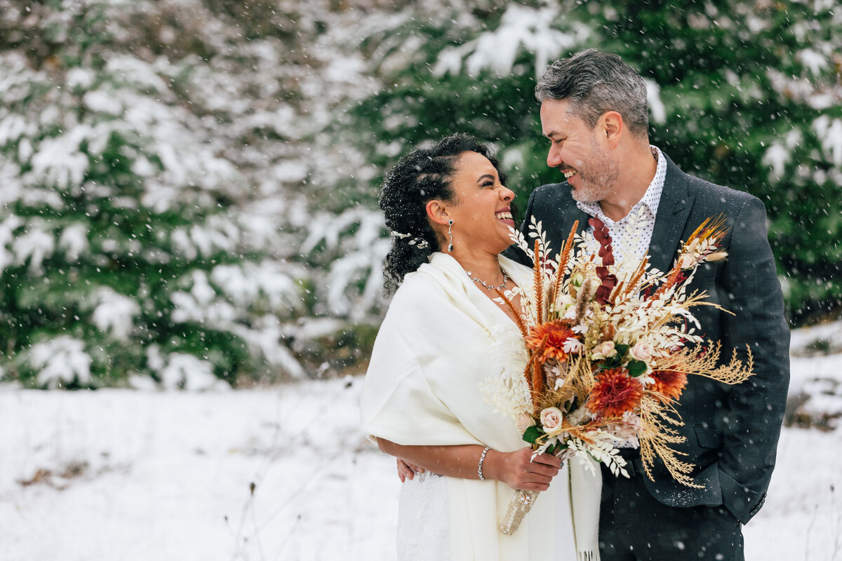 A Puerto Rican bride wearing a cream shawl and holding dried flowers laughs as she looks at her groom in a gray suit jacket and blue polka dot shirt as snow falls among the Evergreen trees at their Columbia River Gorge PNW winter adventure elopement at Government Cove near Hood River, Oregon. | Erica Swantek Photography