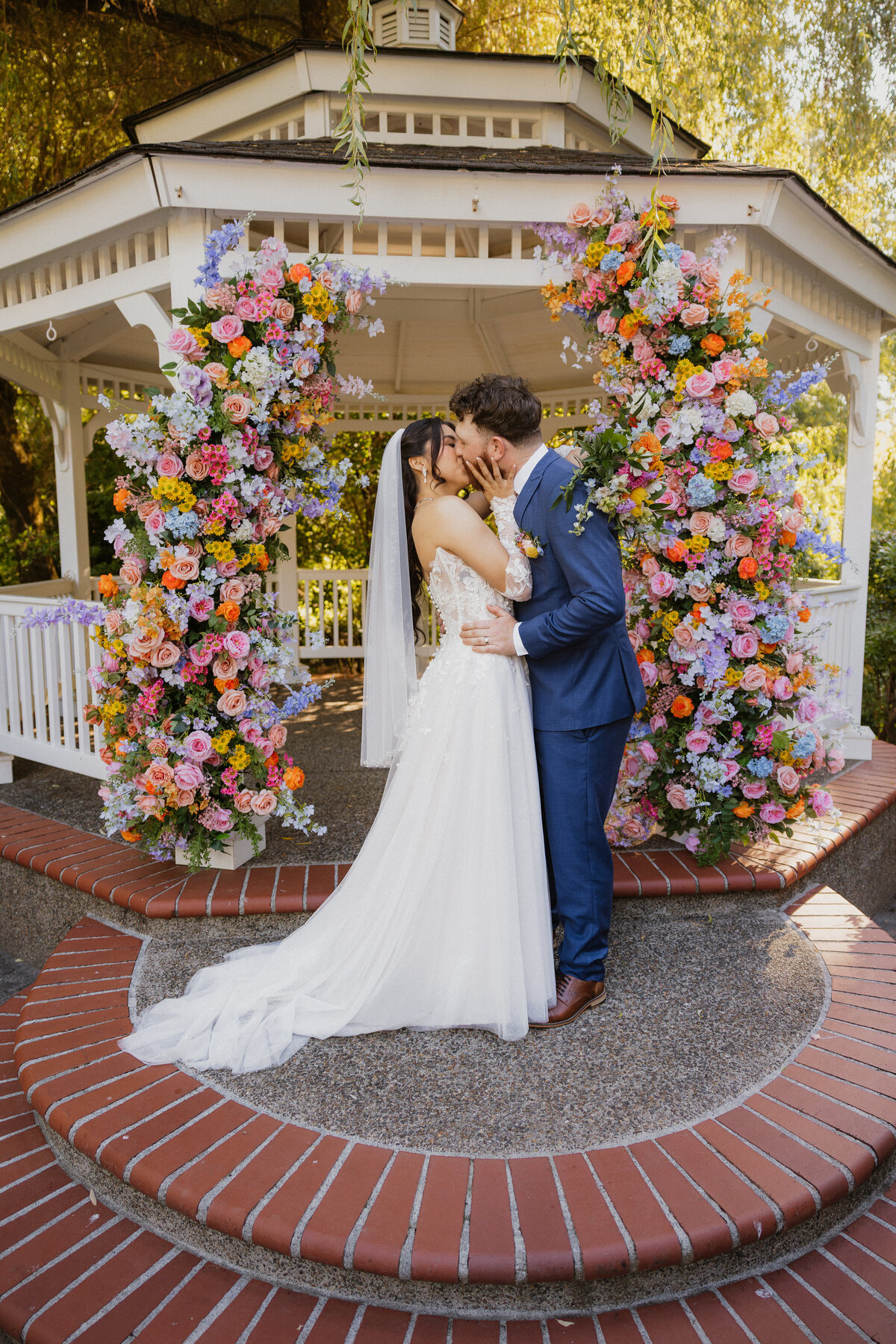 Newlyweds kiss beneath a gazebo covered in colorful flowers.