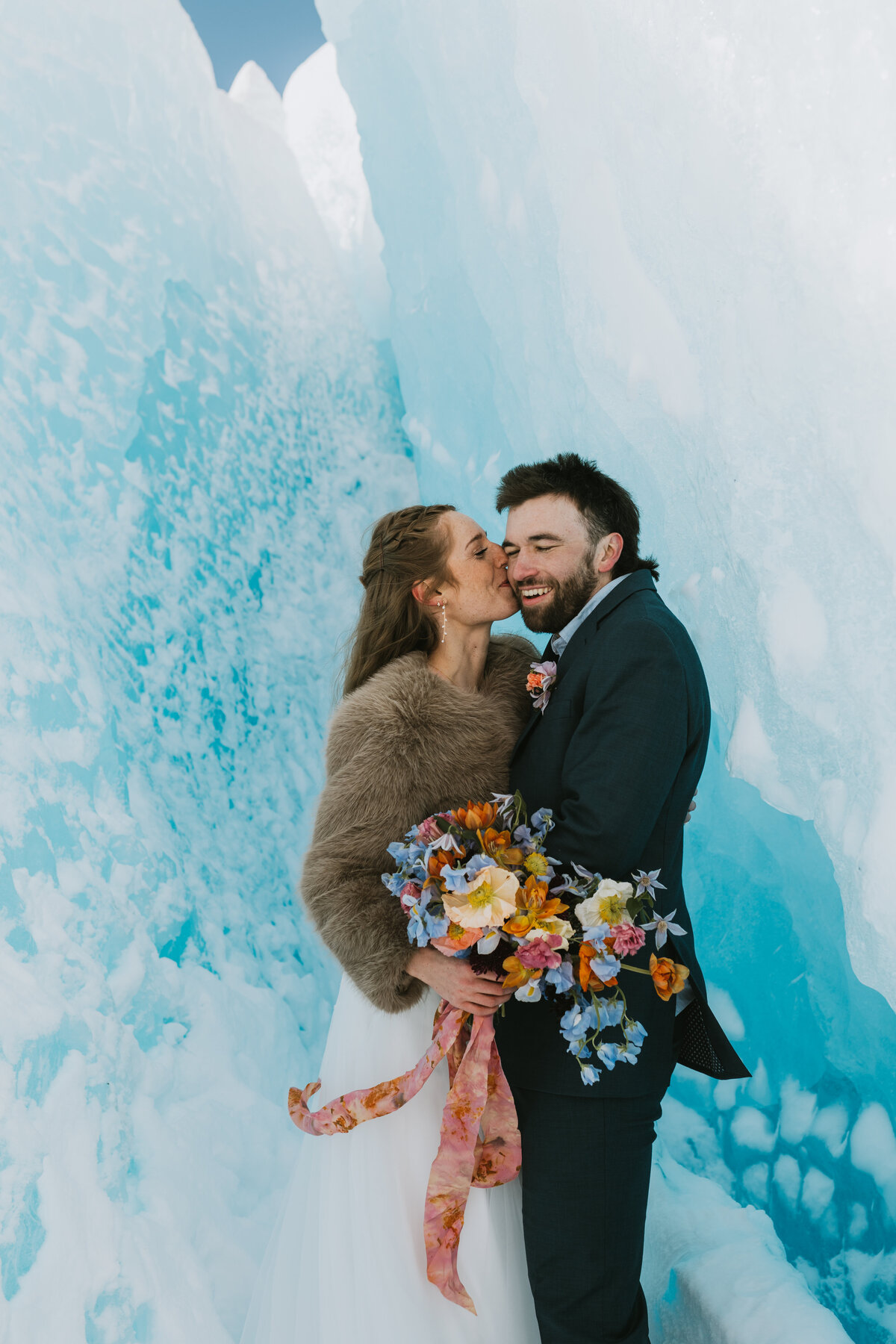 A happy just married couple in their wedding attire snuggling close and kissing in a blue glacier cave with colorful flowers on their elopement day