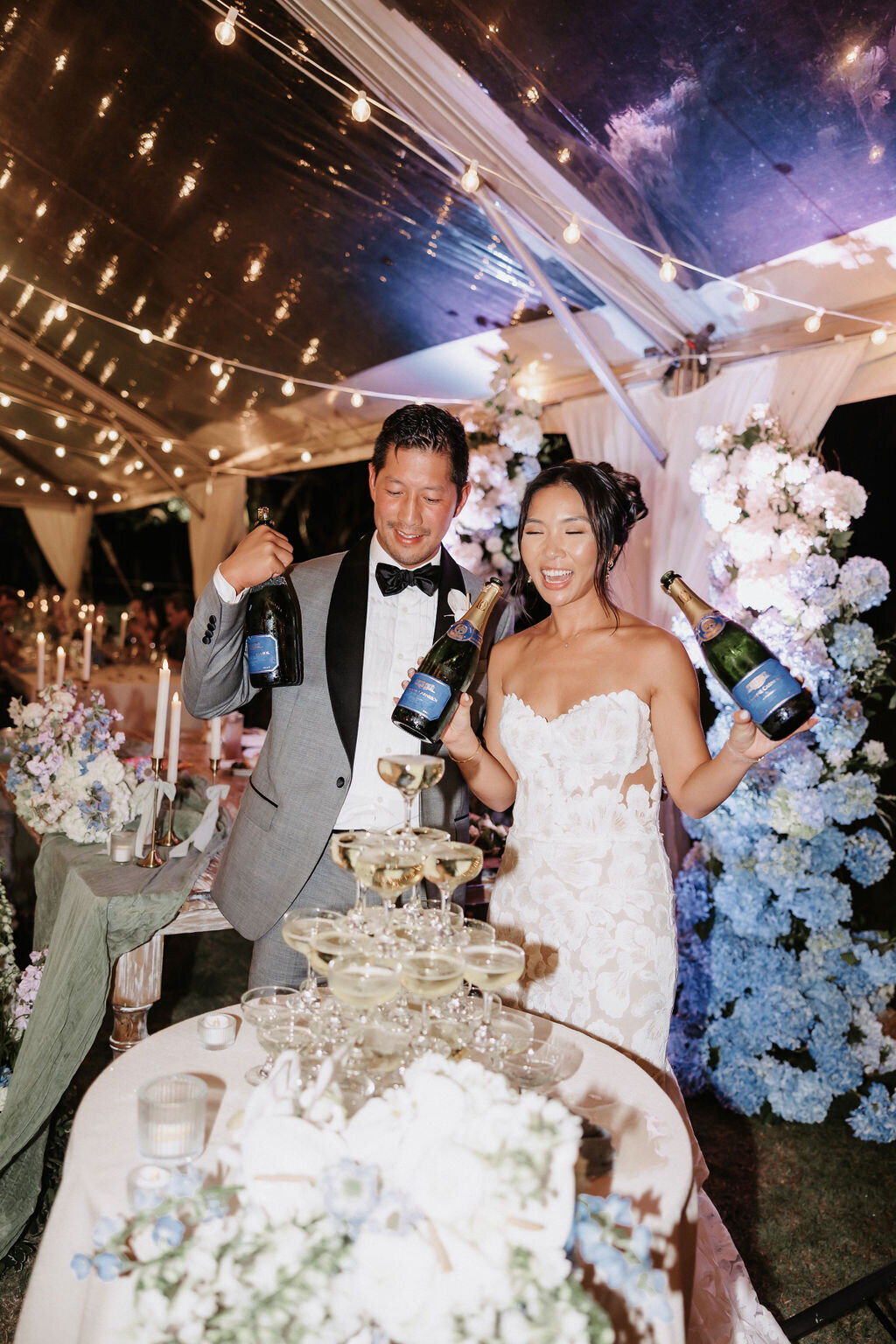 Bride and groom standing behind champagne tower at wedding guest candid photo under clear tent at lanikuhonua