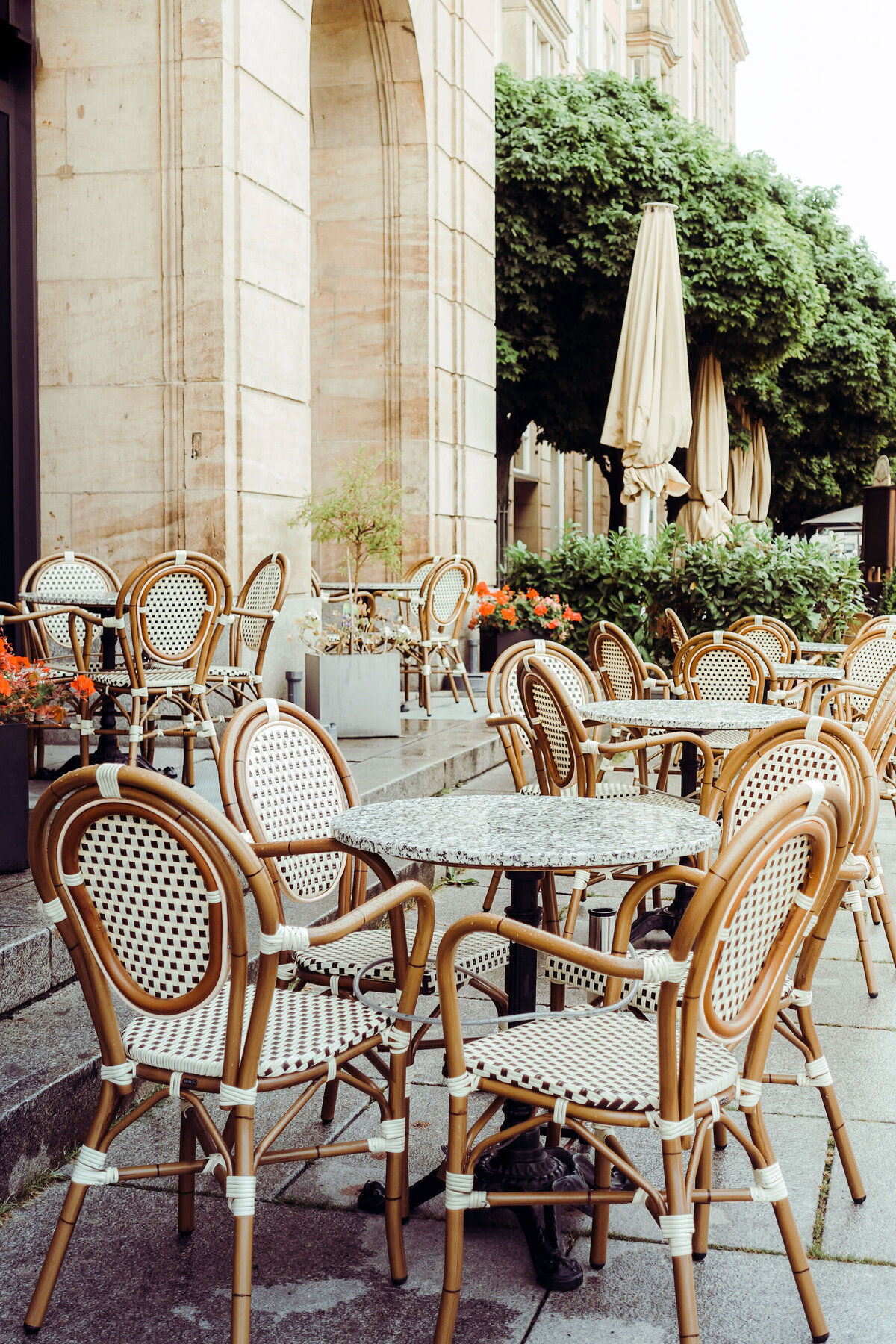 Outdoor cafe with empty wicker chairs and tables on a stone patio. Large trees and beige umbrellas provide shade, creating an ambiance fit for luxury vacations. Orange flowers in planters add color to the scene beside a building adorned with tall arches and stone walls.