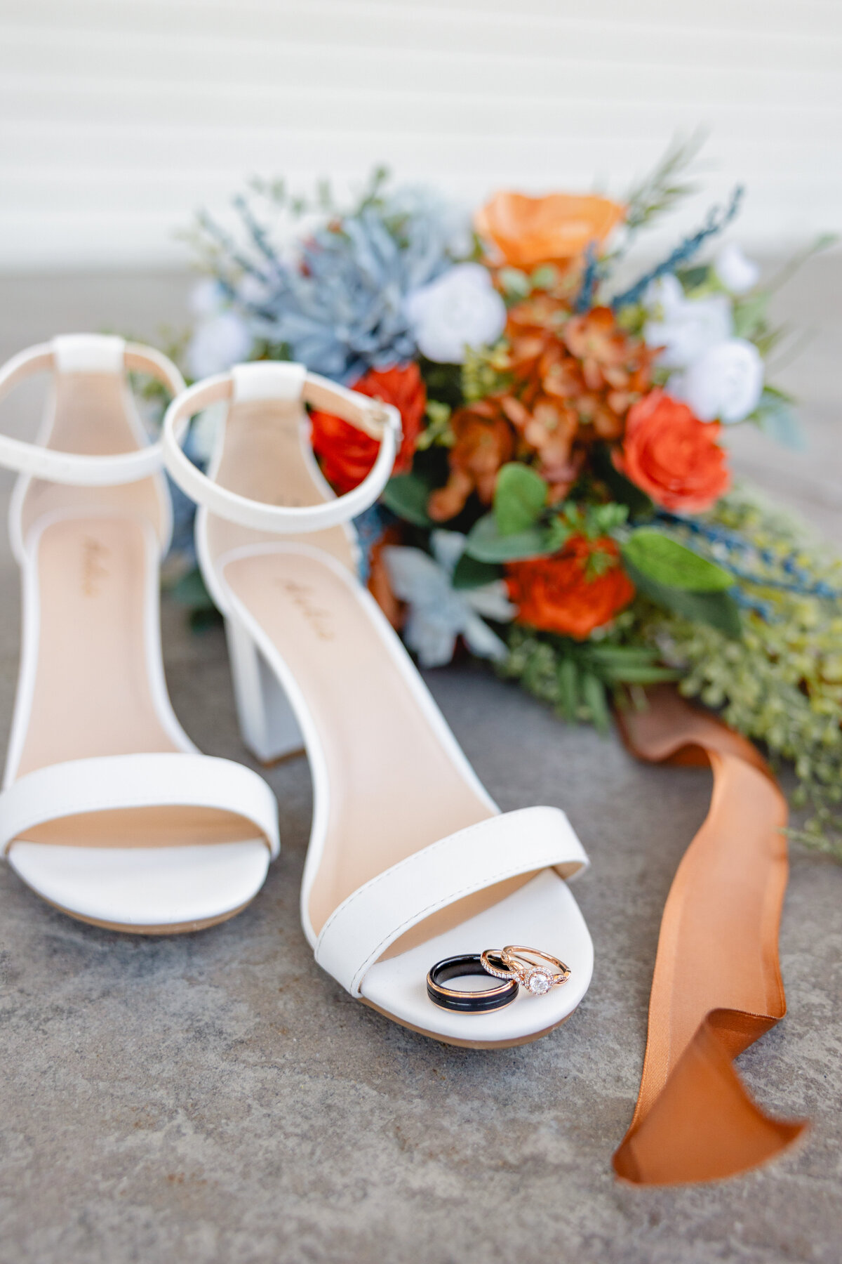 A pair of white bridal heels with wedding rings placed on the toe of one shoe, set beside a colorful bouquet with orange, blue, and white flowers, captured by Cassie Lee Photography.